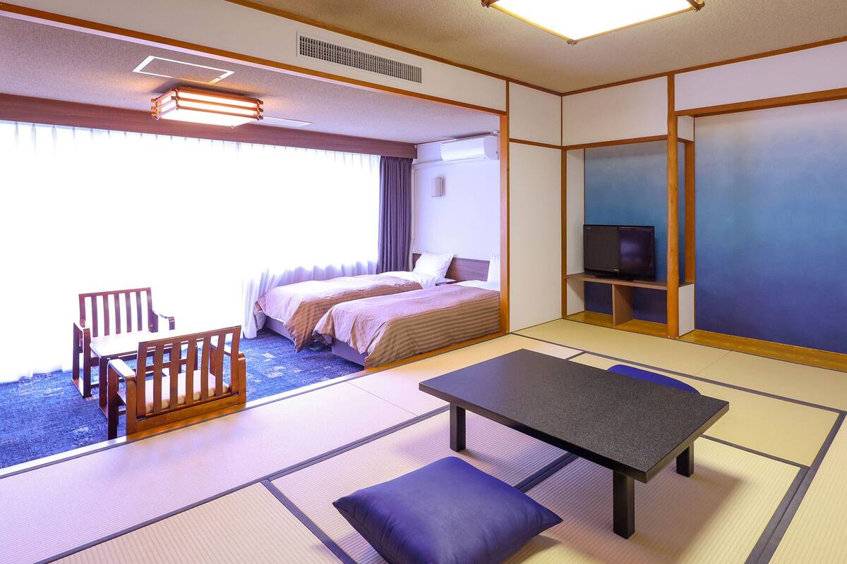 Ett traditionellt japanskt hotellrum med tatamimattor på golvet, en säng, en TV och en liten sittgrupp.