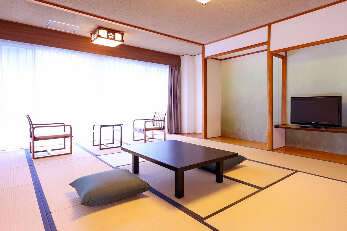 Ett traditionellt japanskt rum med tatamimattor på golvet, ett lågt bord, två stolar, en tv på en hylla och skjutdörrar.