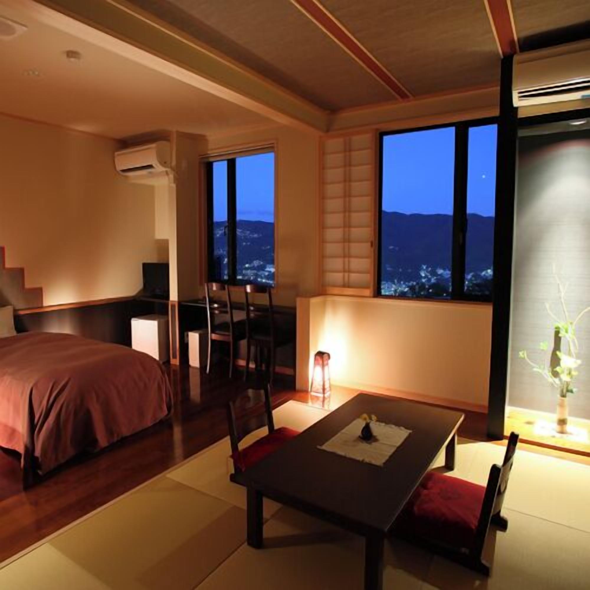 A hotel room with a bed, a dining table with chairs, a view of the city at dusk, and a traditional Japanese sliding door.