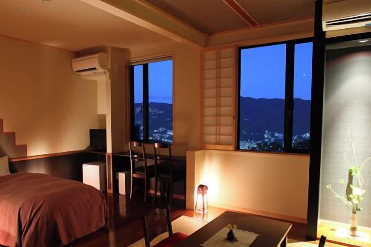 A hotel room with a bed, a dining table with chairs, a view of the city at dusk, and a traditional Japanese sliding door.