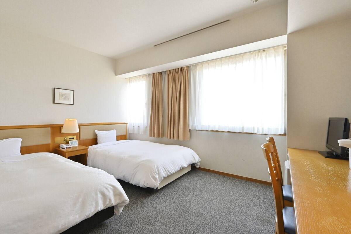 Une chambre d’hôtel avec deux lits, un bureau avec une chaise, une télévision et une fenêtre avec des rideaux.