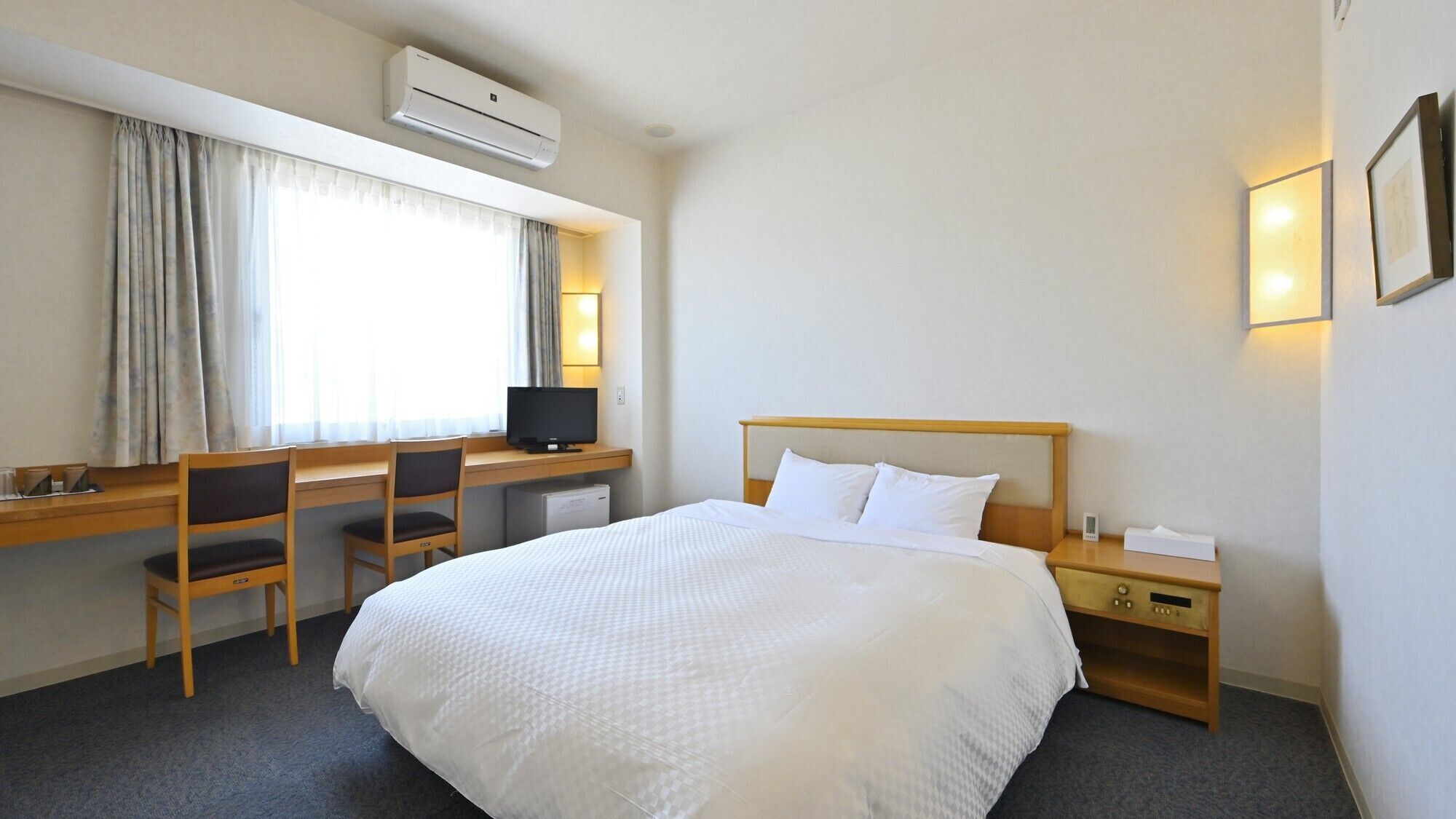 A hotel room with a bed, desk, chair, TV, and bedside table.