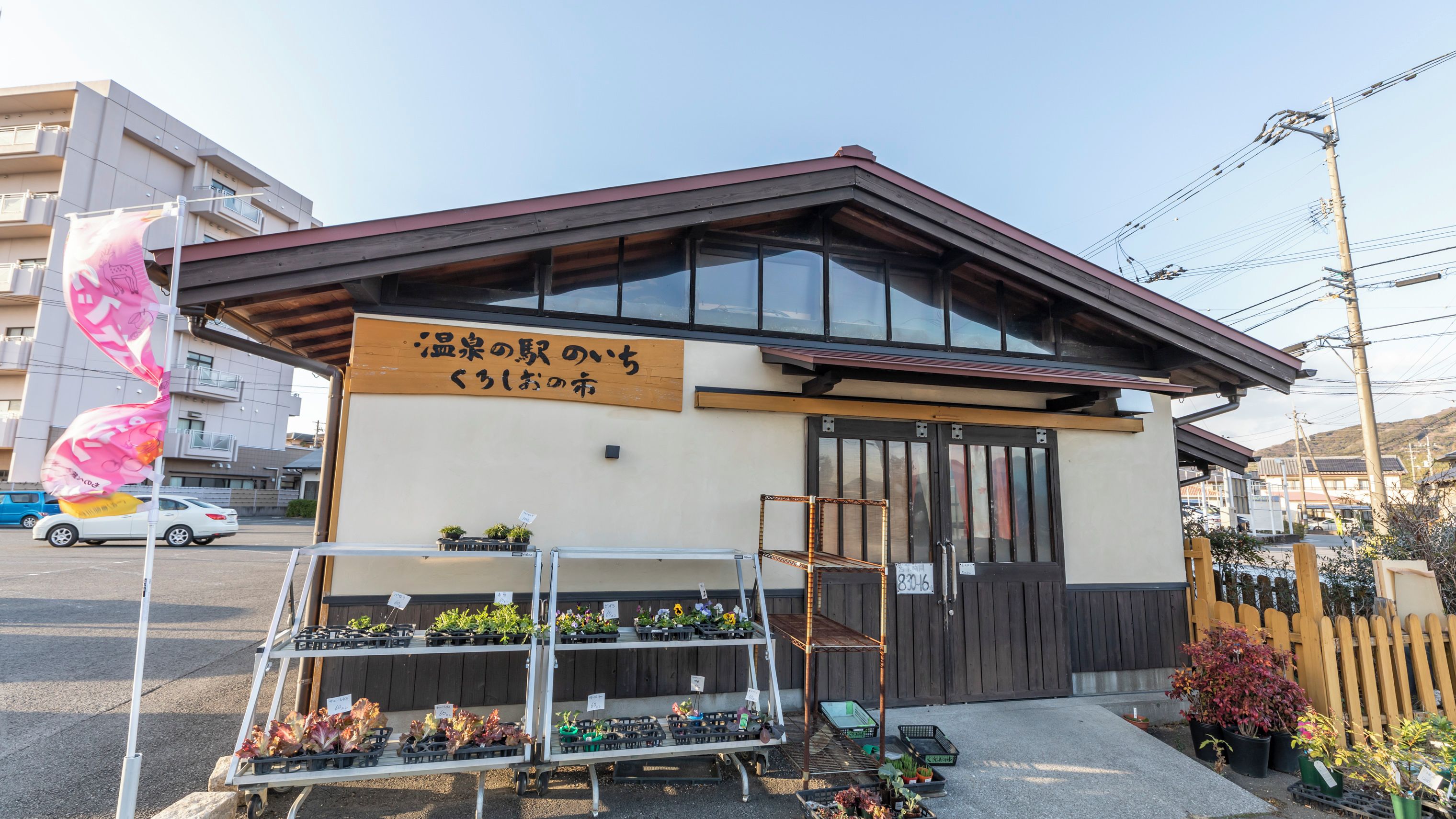 A building with a sign in Japanese, a storefront with potted plants, and a wooden fence in the background.