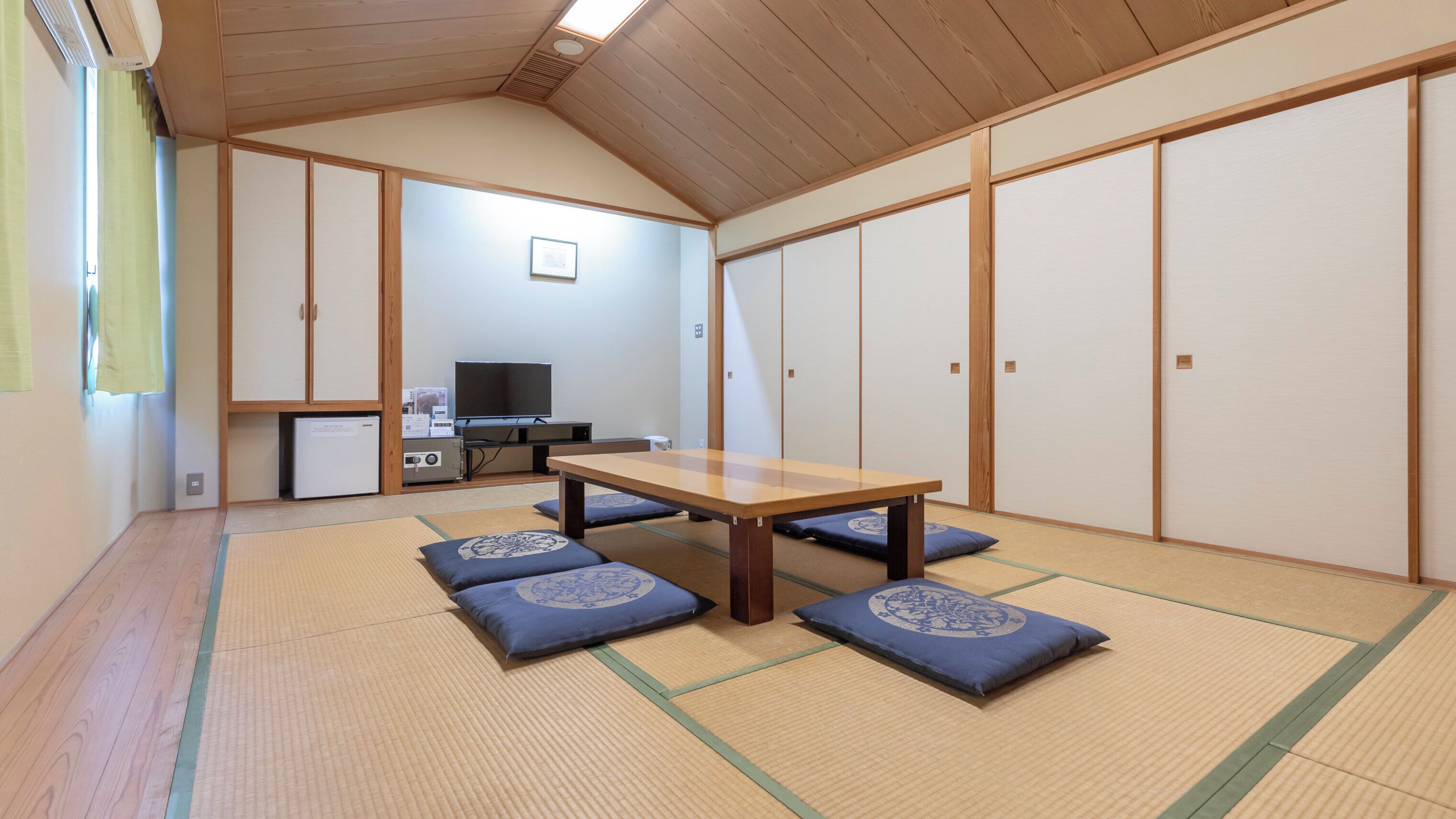 A traditional Japanese room with tatami flooring, a low table, cushions, a television, and a refrigerator.