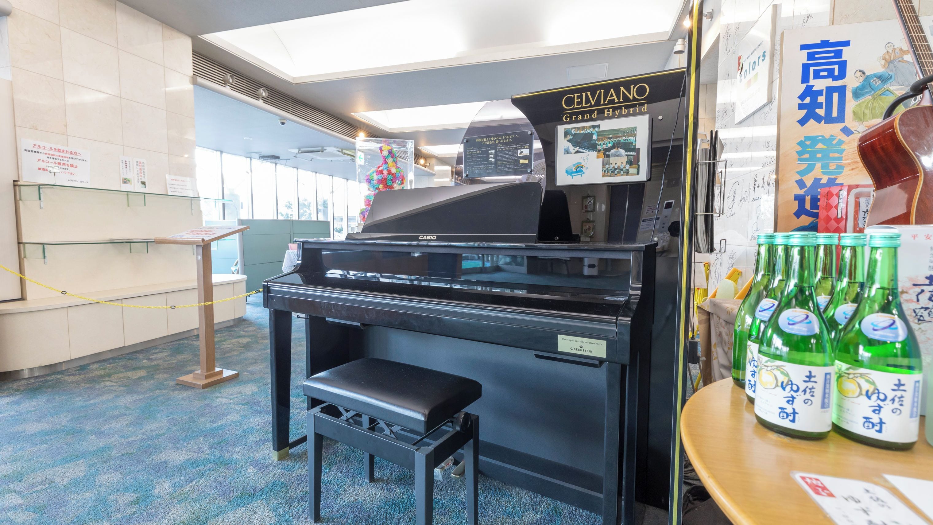 A piano with a Casio Grand Hybrid label, a stool, and a display of green bottles with Japanese text.