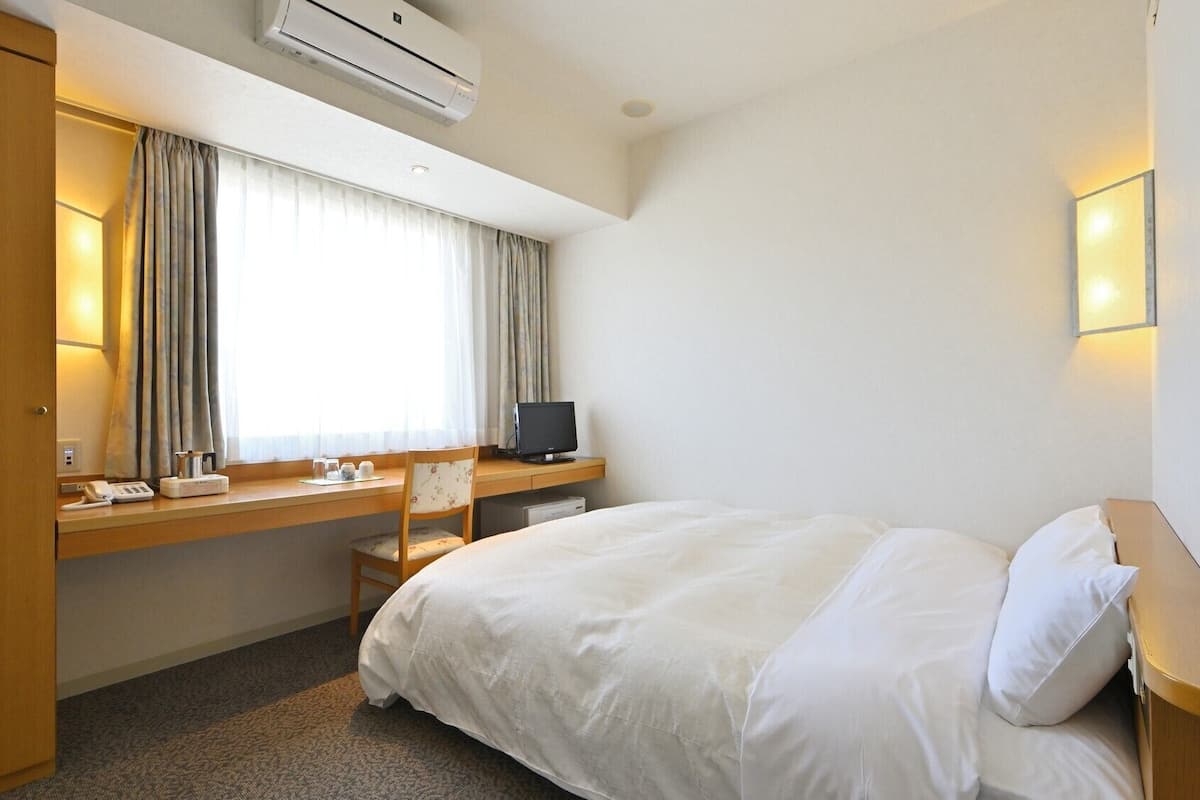 Une chambre d’hôtel comprenant un lit, un bureau avec un ordinateur, une chaise, une fenêtre avec des rideaux, un climatiseur et une table de chevet.
