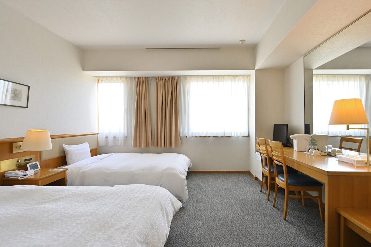 Habitación de hotel con dos camas, un escritorio, una silla, un espejo, una lámpara y un reloj.