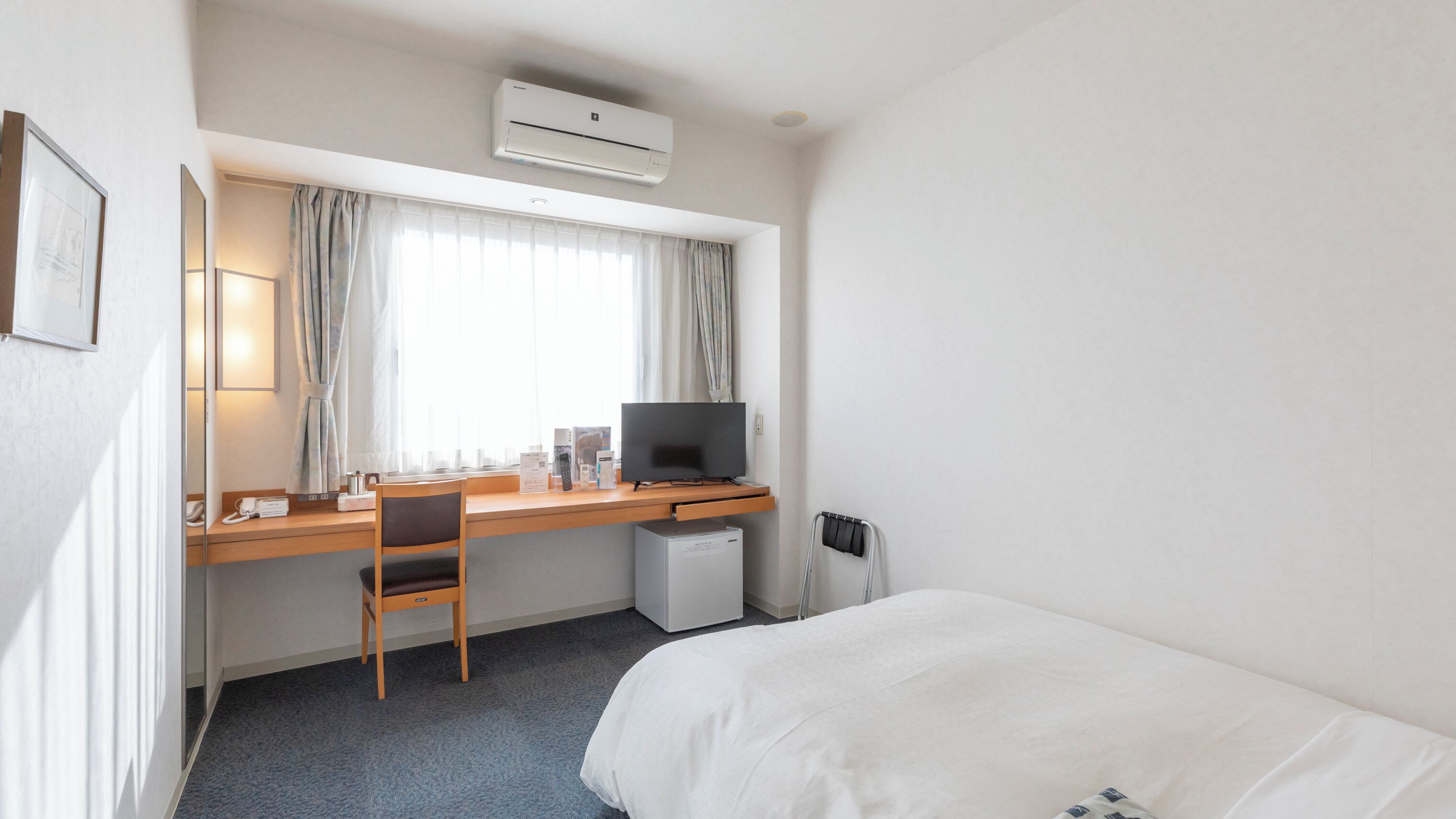 Habitación de hotel con cama, escritorio con computadora, silla, refrigerador pequeño y ventana con cortinas.