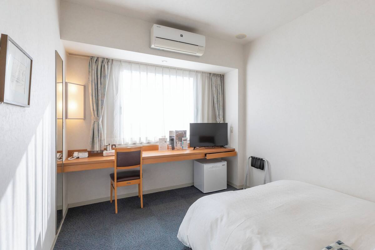 Une chambre d’hôtel comprenant un lit, un bureau avec un ordinateur, une chaise, un petit réfrigérateur et une fenêtre avec des rideaux.