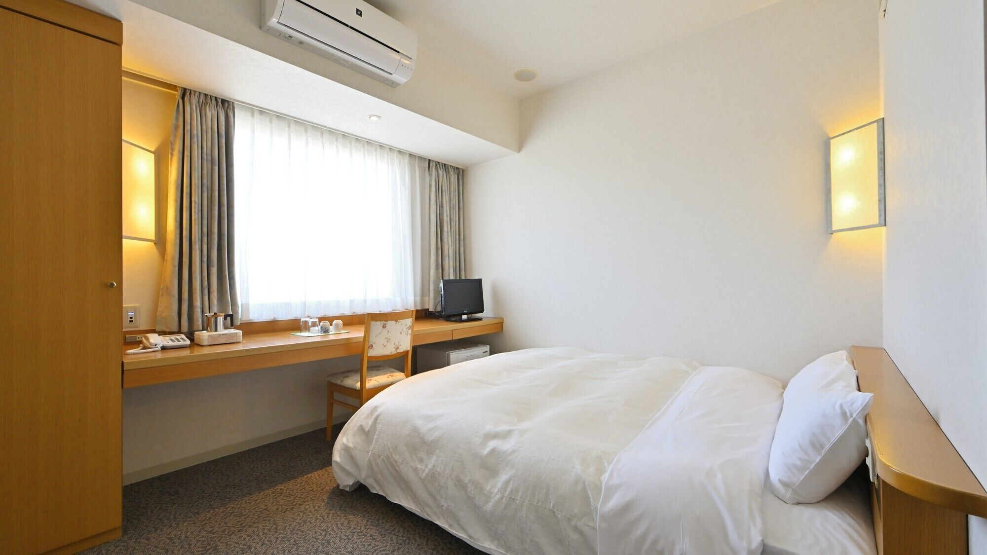 Una habitación de hotel con cama, escritorio con computadora, silla, ventana con cortinas, unidad de aire acondicionado y mesita de noche con lámpara.