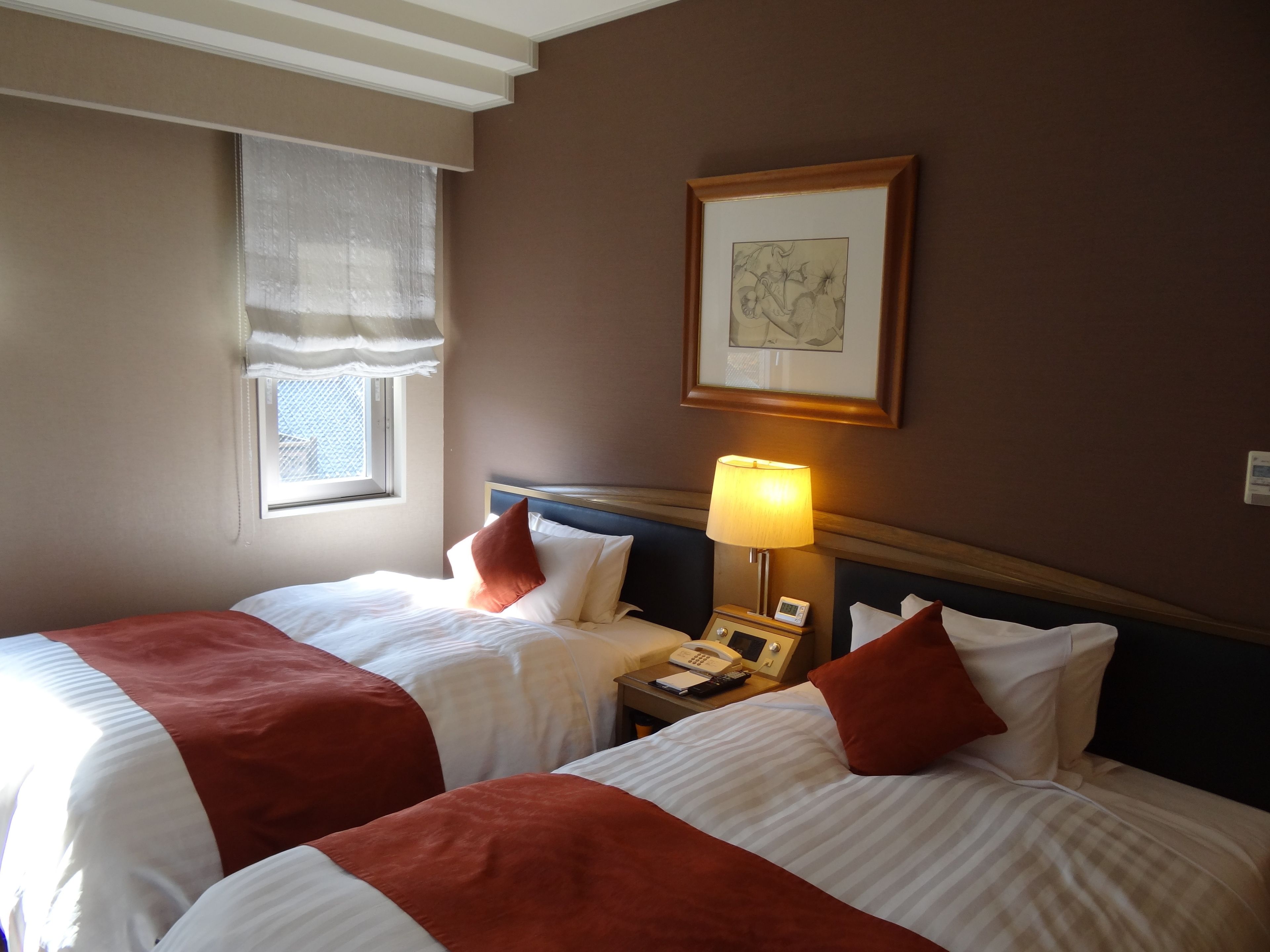 A hotel room with two beds, a bedside table with a lamp, a framed picture on the wall, and a window with a curtain.