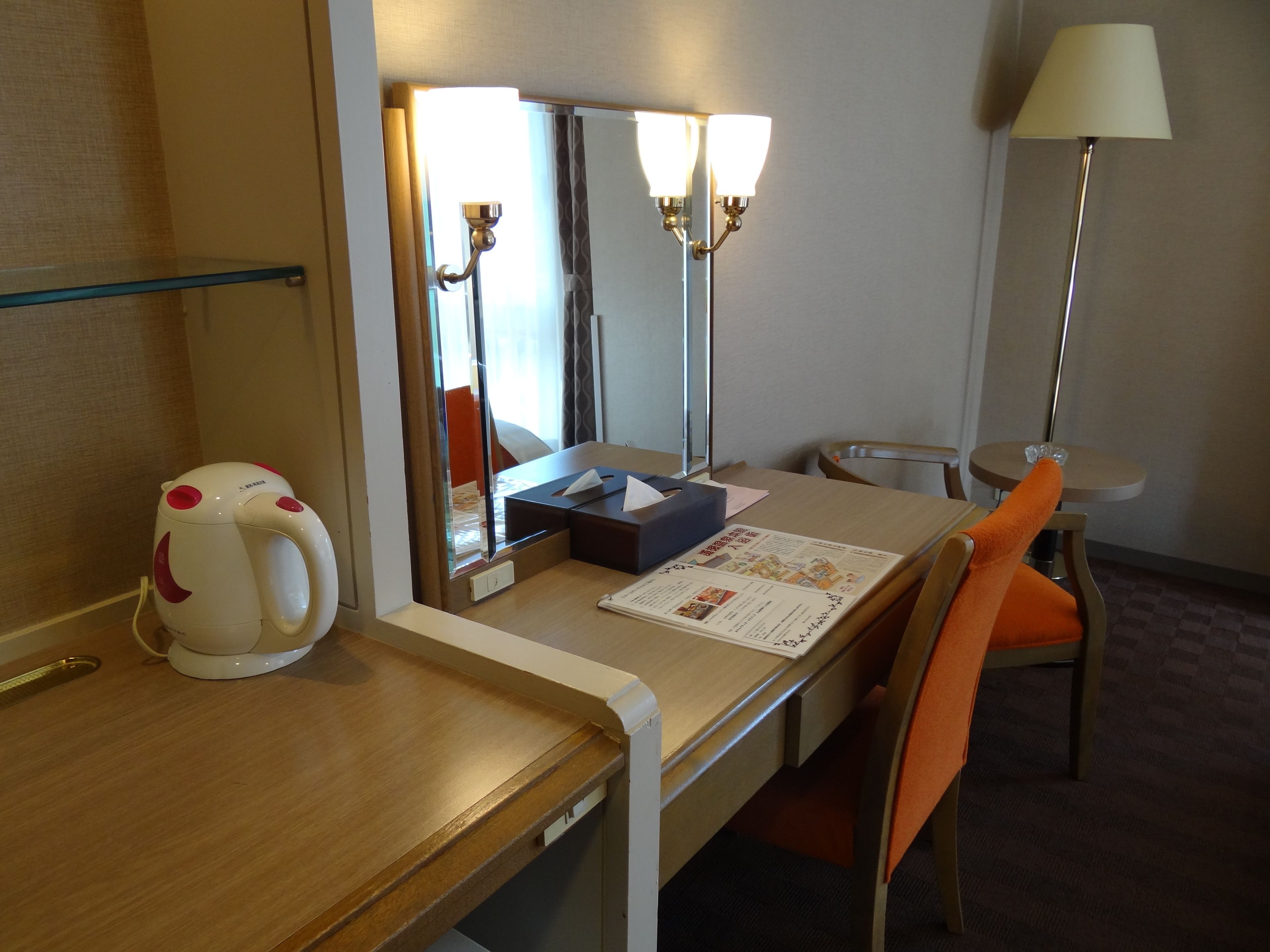 Een hotelkamer met een houten bureau, een waterkoker, een spiegel, een lamp en een stoel.