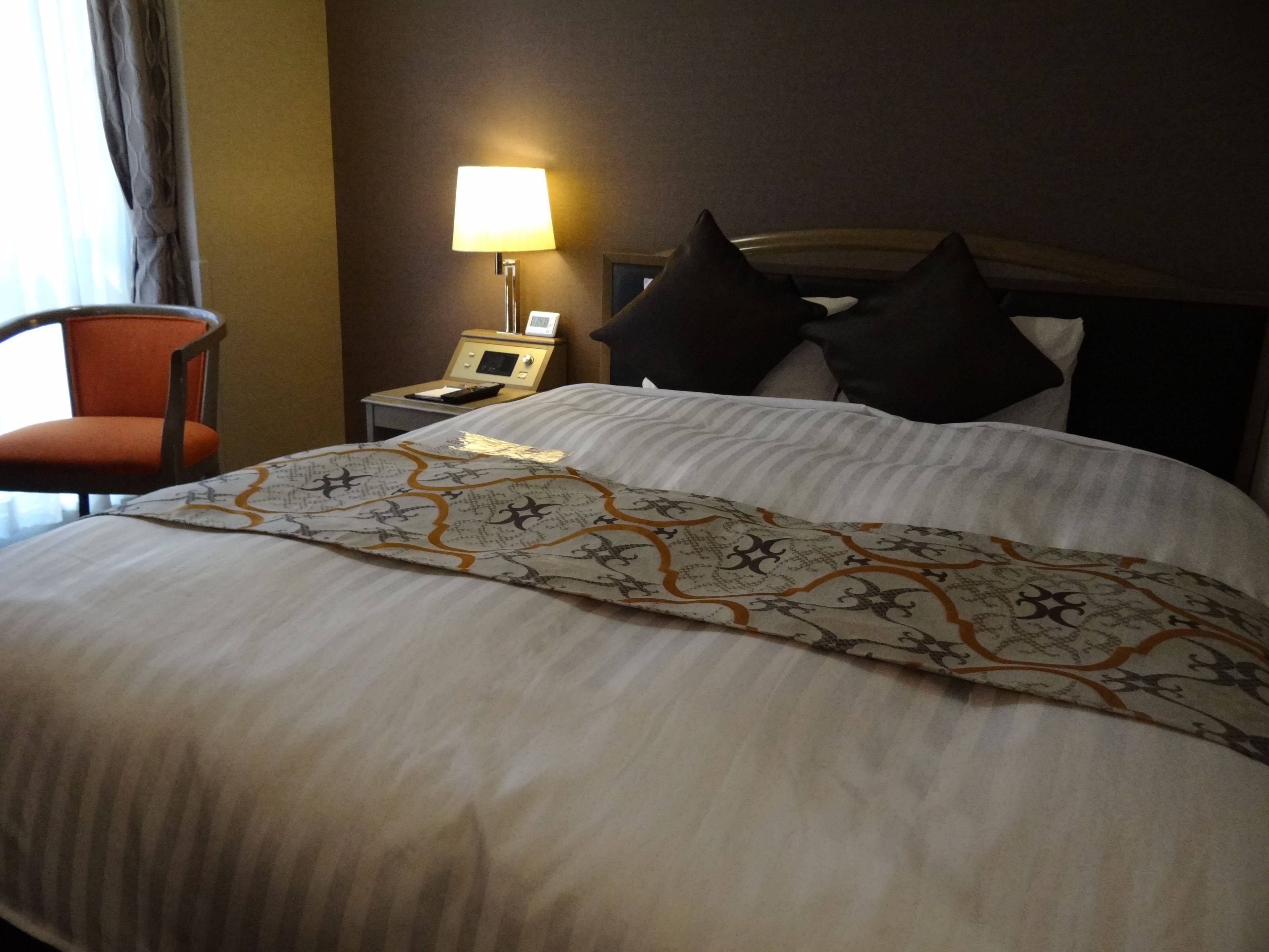 A hotel room with a neatly made bed, a bedside table with a lamp, and a chair.
