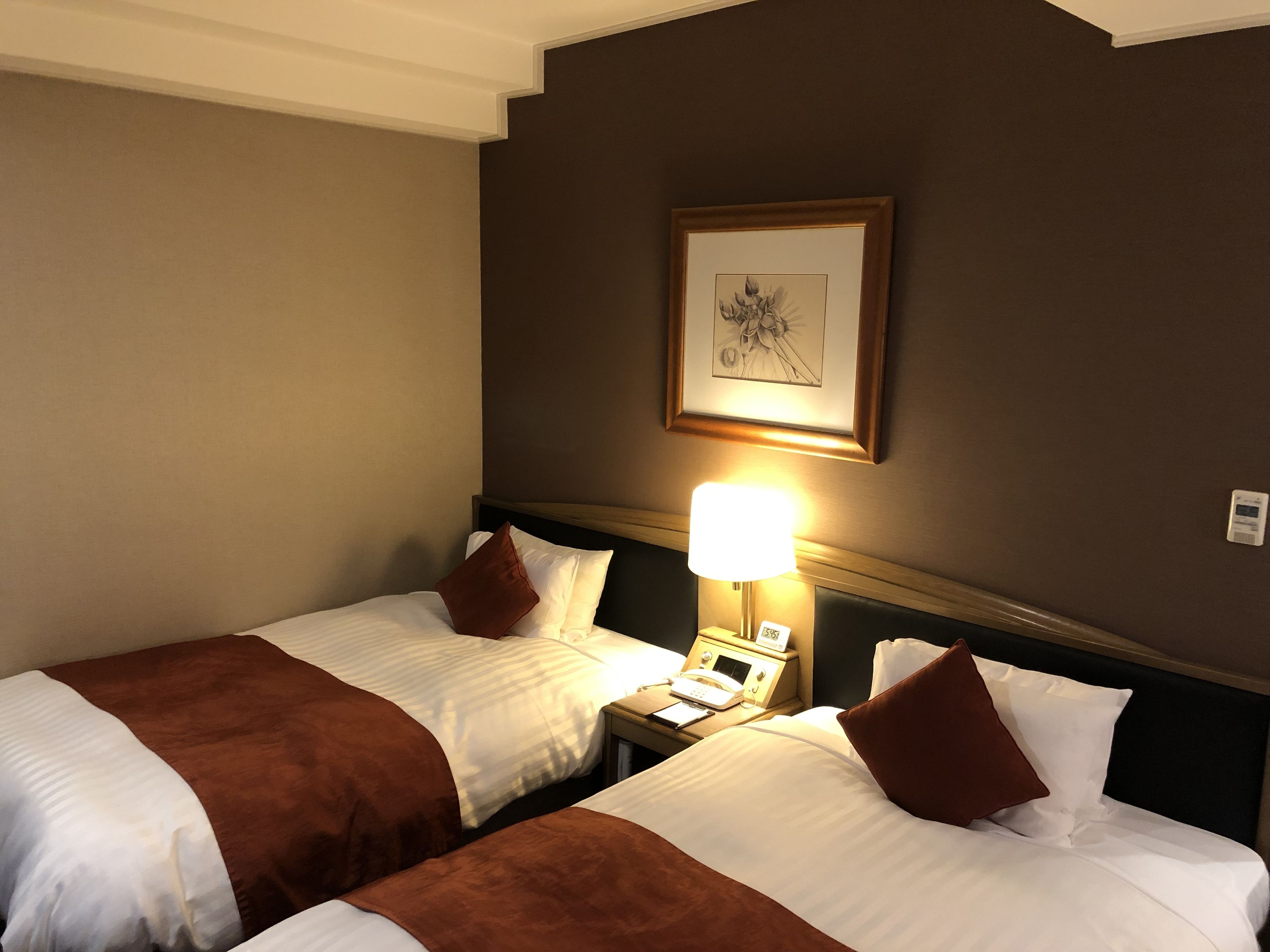 A hotel room with two beds, a bedside table with a lamp, and a framed picture on the wall.