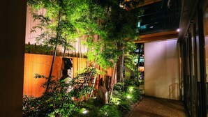 A narrow brick pathway flanked by lush greenery and wooden fences, leading to a building with multiple balconies. - Hotel Patio Dogo (Matsuyama)