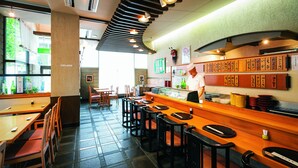 A restaurant interior with wooden tables and black bar stools, a counter with a menu, and a view of the outdoors. - Hotel Patio Dogo (Matsuyama)