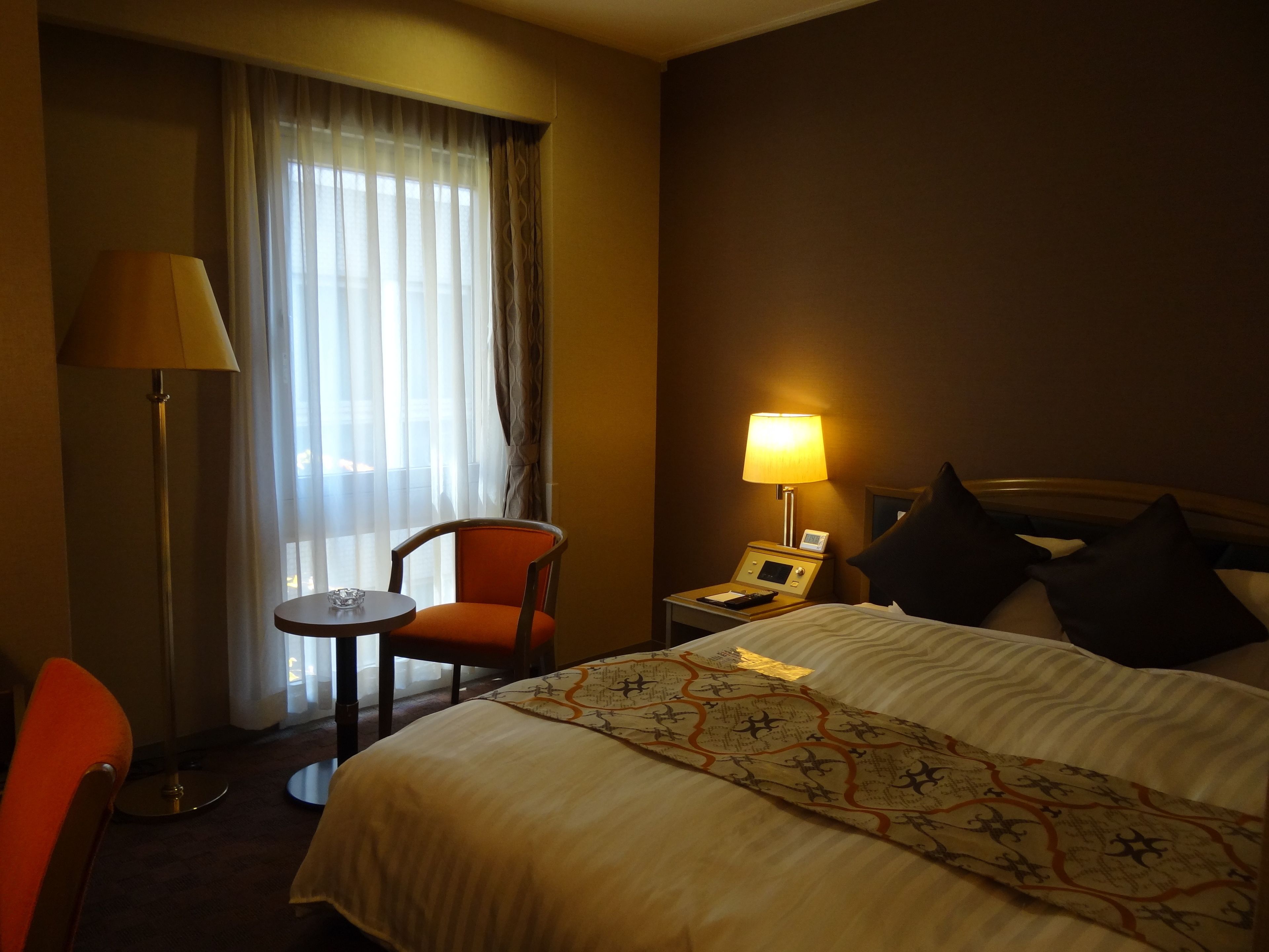 Ein Hotelzimmer mit Bett, Stuhl, Tisch, Lampe und einem Fenster mit Vorhängen.