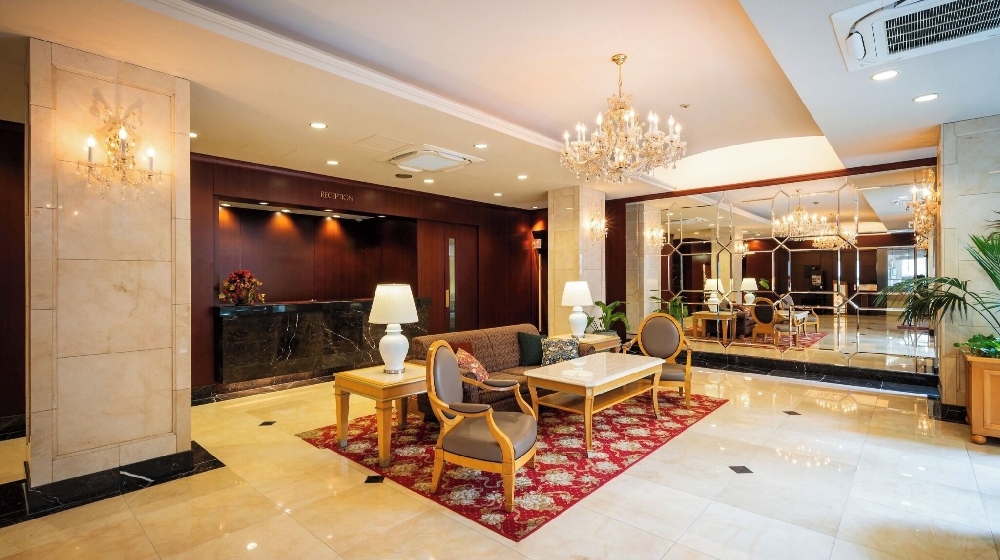 A hotel reception area with a reception desk, seating, a chandelier, and a mirror reflecting the interior.
