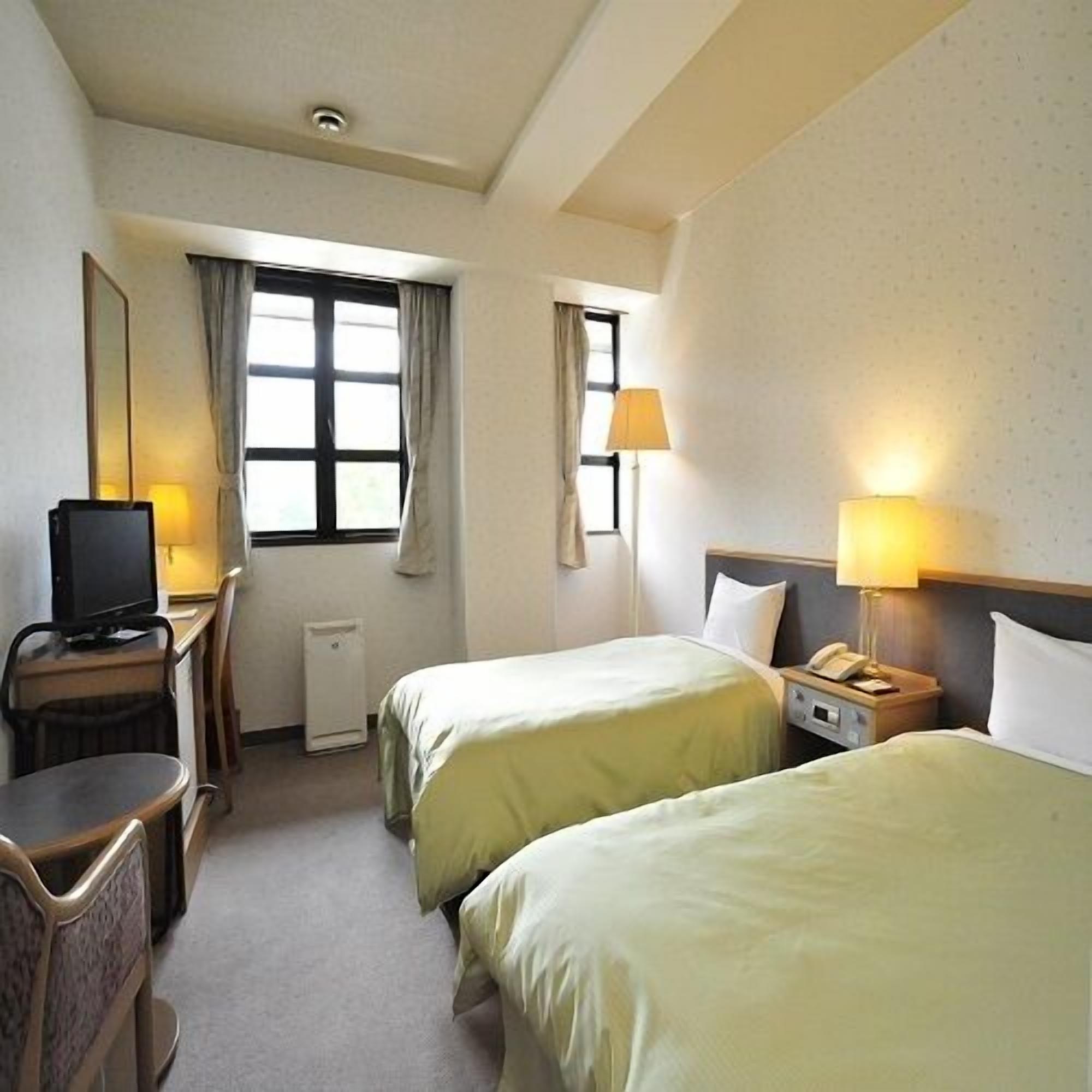 A hotel room with two beds, a desk with a television, a lamp, and a window.