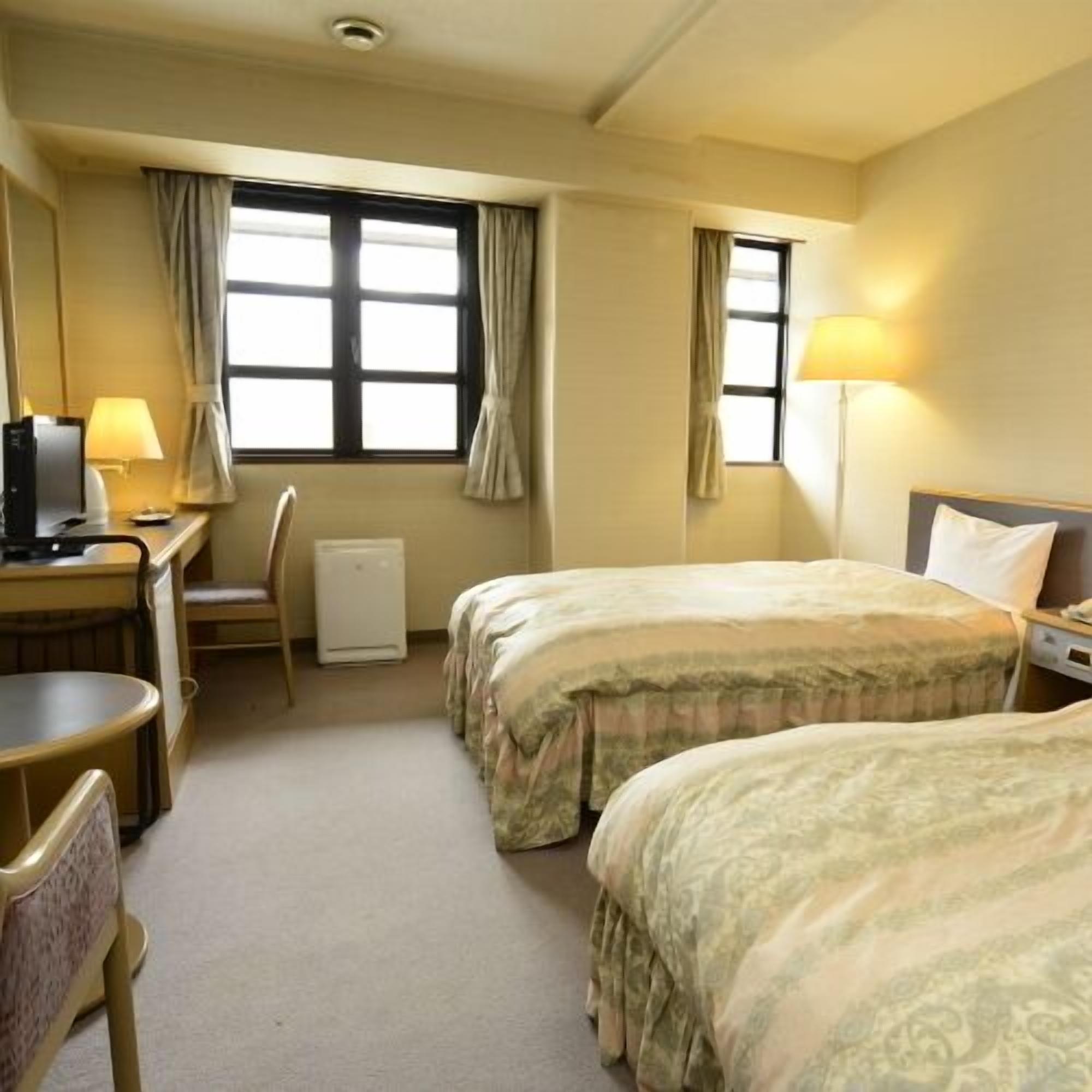 Habitación de hotel con dos camas, un escritorio con computadora, una silla y una ventana con cortinas.