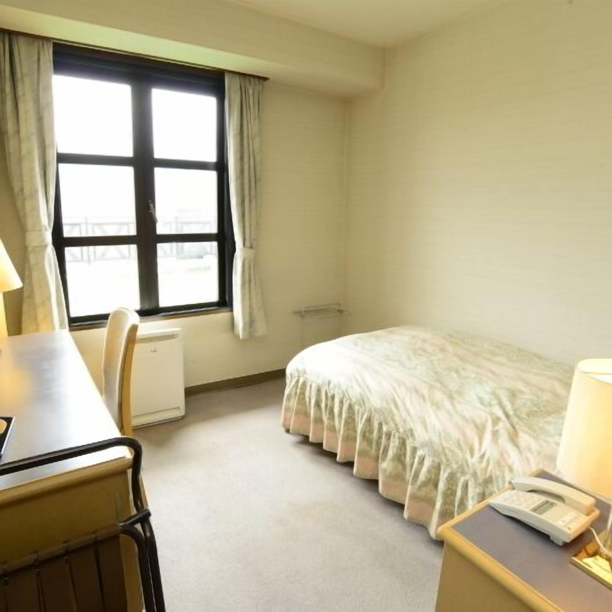 Habitación de hotel con cama, escritorio, silla, ventana con cortinas y teléfono junto a la cama.