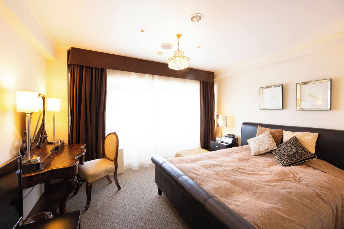 Habitación de hotel con cama, escritorio, silla y un ventanal con cortinas.