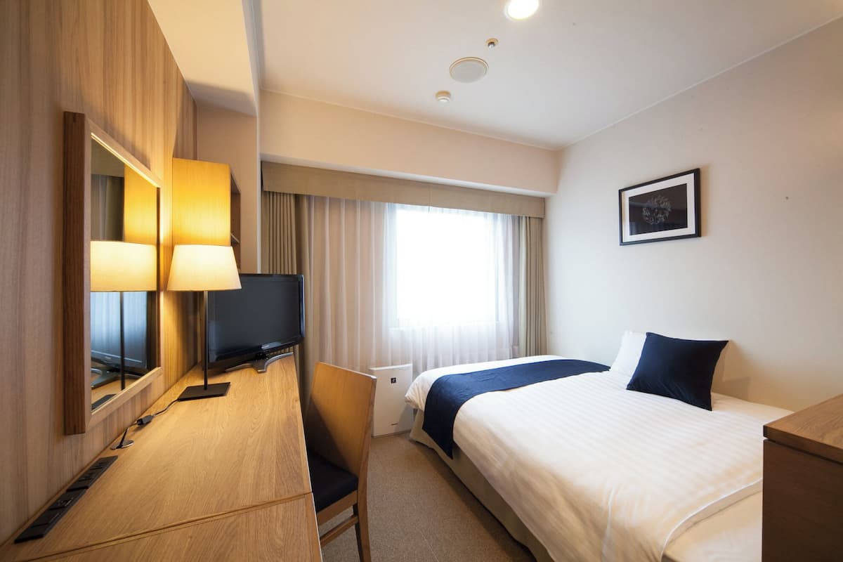 Una habitación de hotel con una cama, un escritorio con computadora y lámpara, una silla, una ventana con cortinas y un cuadro en la pared.