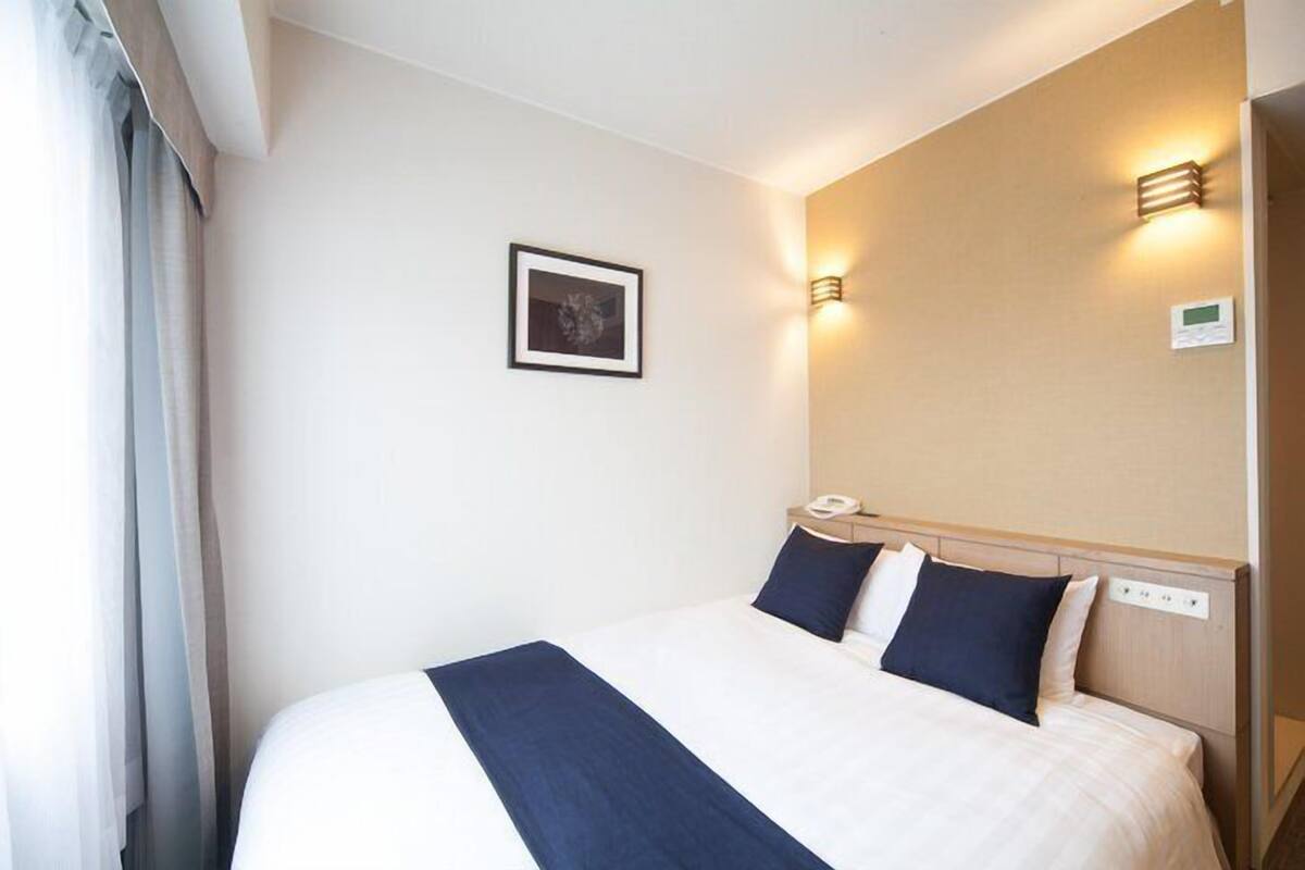 Una habitación de hotel con una cama grande, una mesita de noche con lámpara, un cuadro enmarcado en la pared y un termostato en la pared.