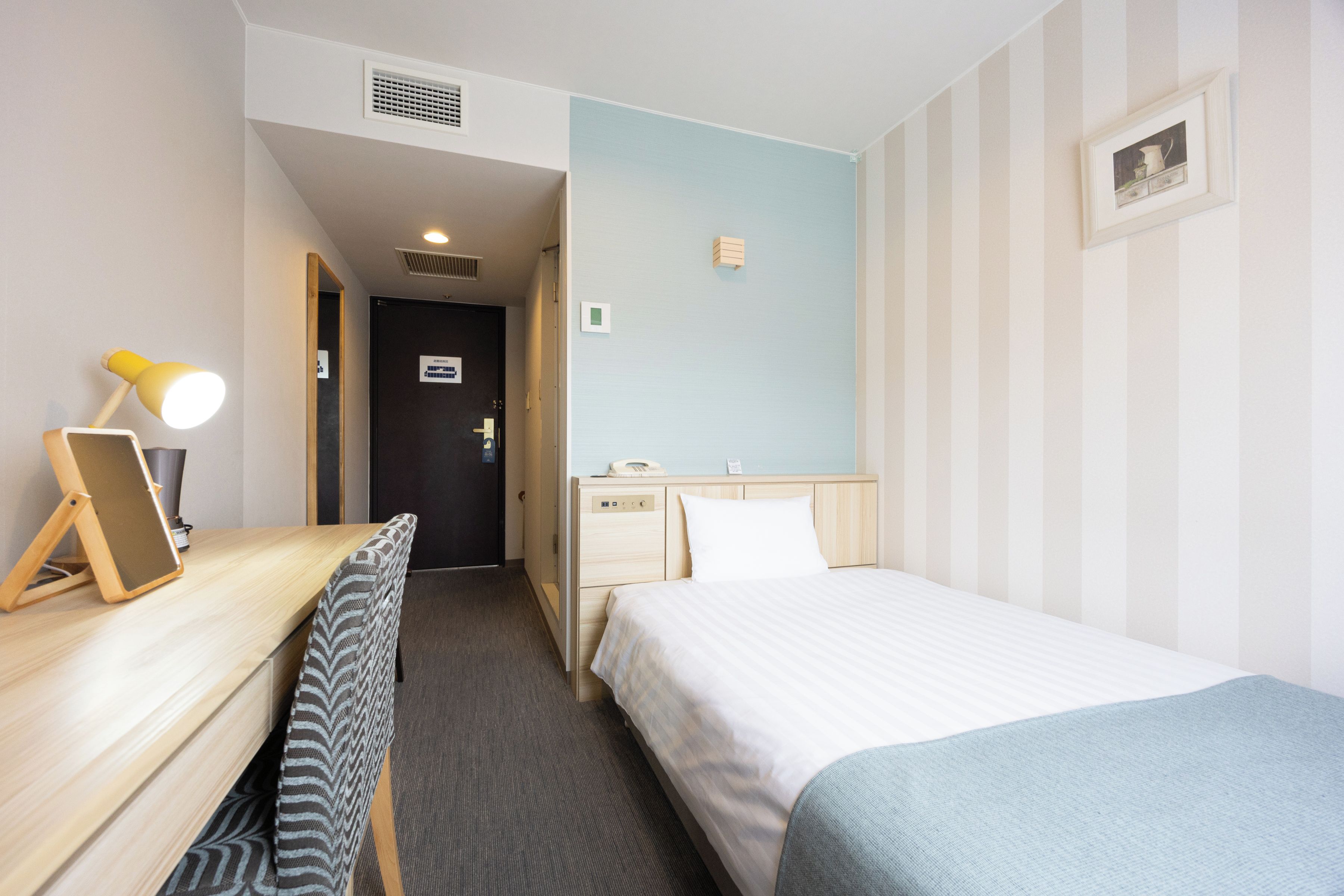 A hotel room with a bed, a desk with a lamp, a bedside table, a mirror, and a small bathroom area.