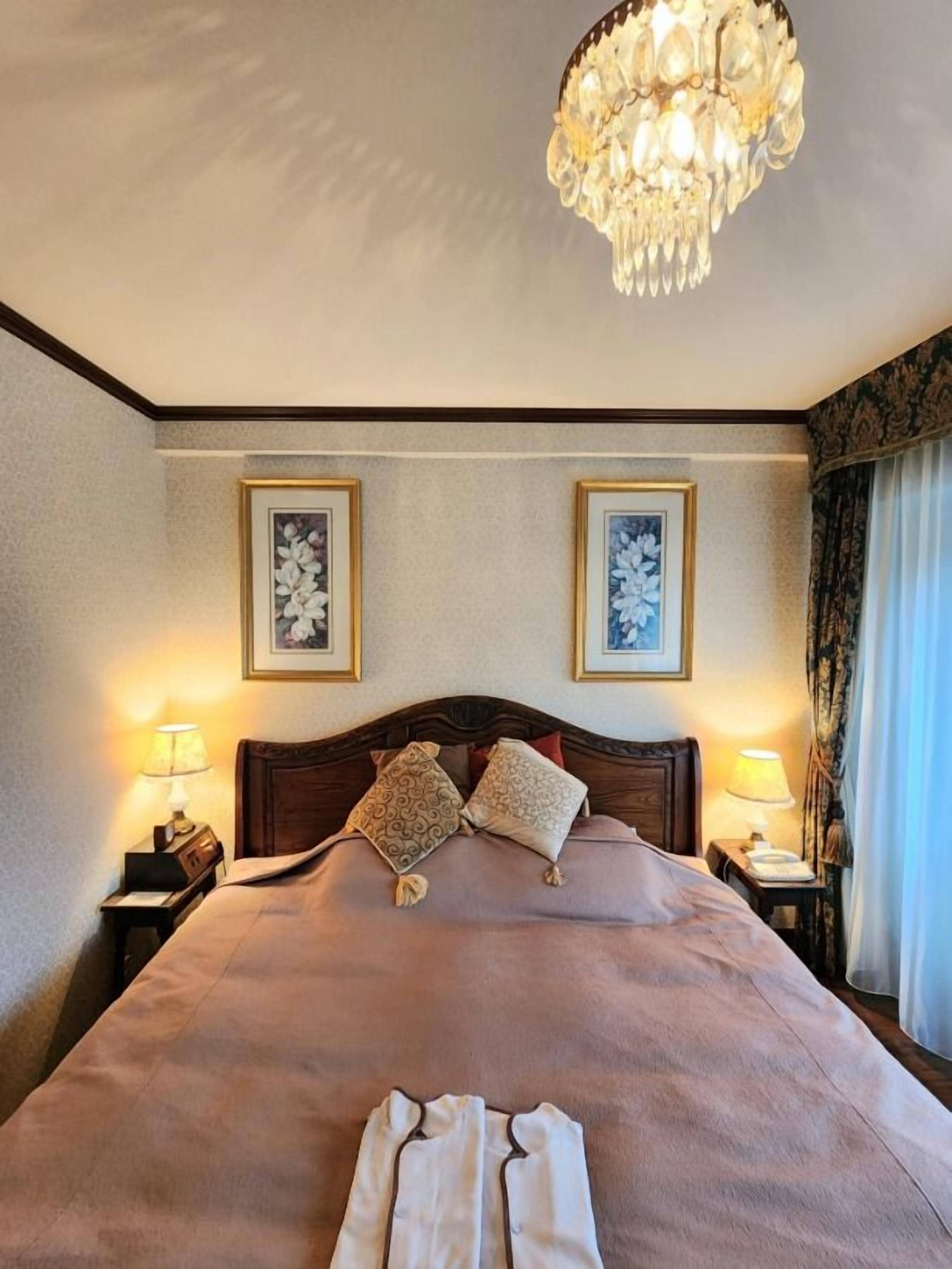 A hotel room with a large bed, two bedside lamps, a chandelier, and framed artwork on the walls.