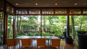 Amplia habitación con un gran comedor y sillas, ventanas amplias que ofrecen vista a un jardín exuberante y árboles.