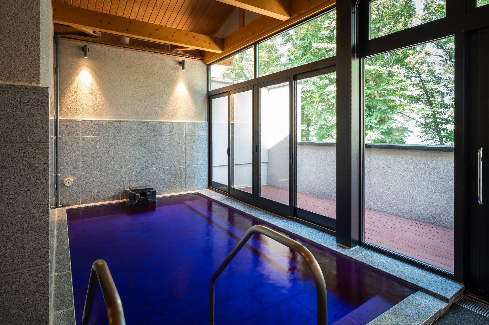 An indoor swimming pool with clear glass walls, a wooden ceiling, and a view of trees outside.