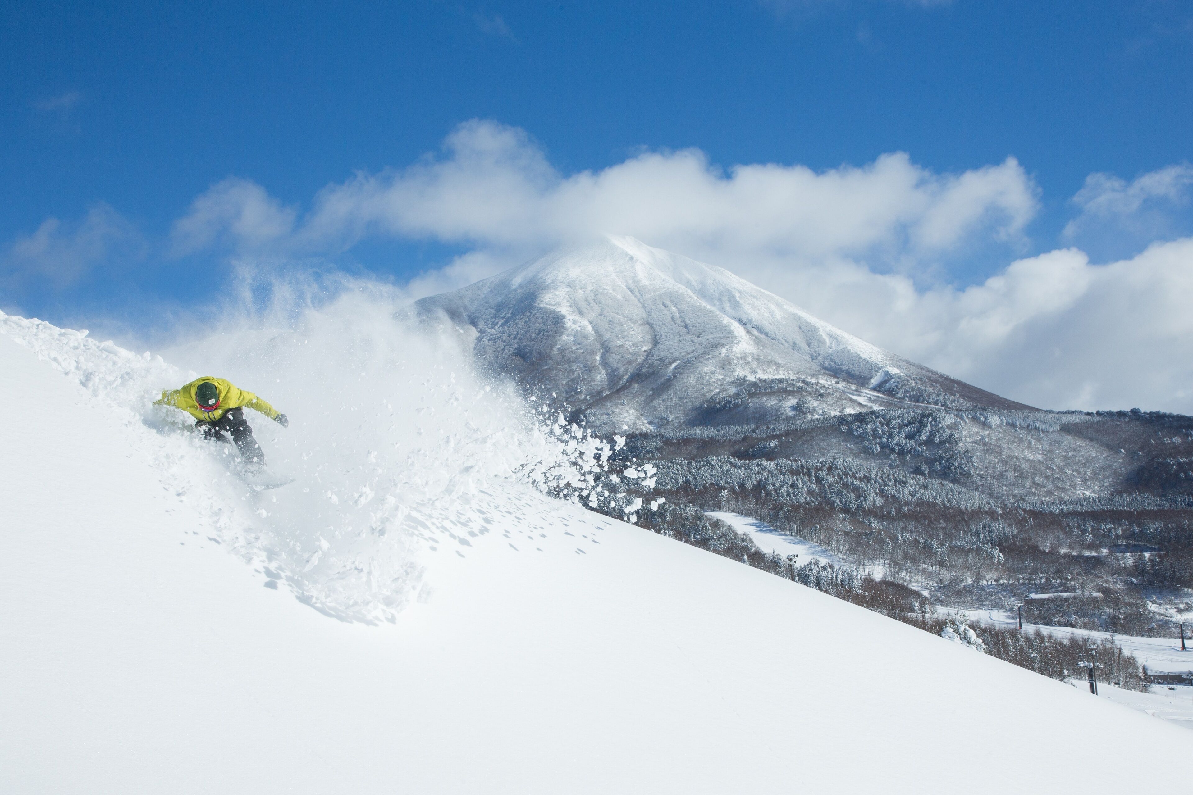 A snowboarder in a yellow jacket on a snowy slope with a mountain in the background.