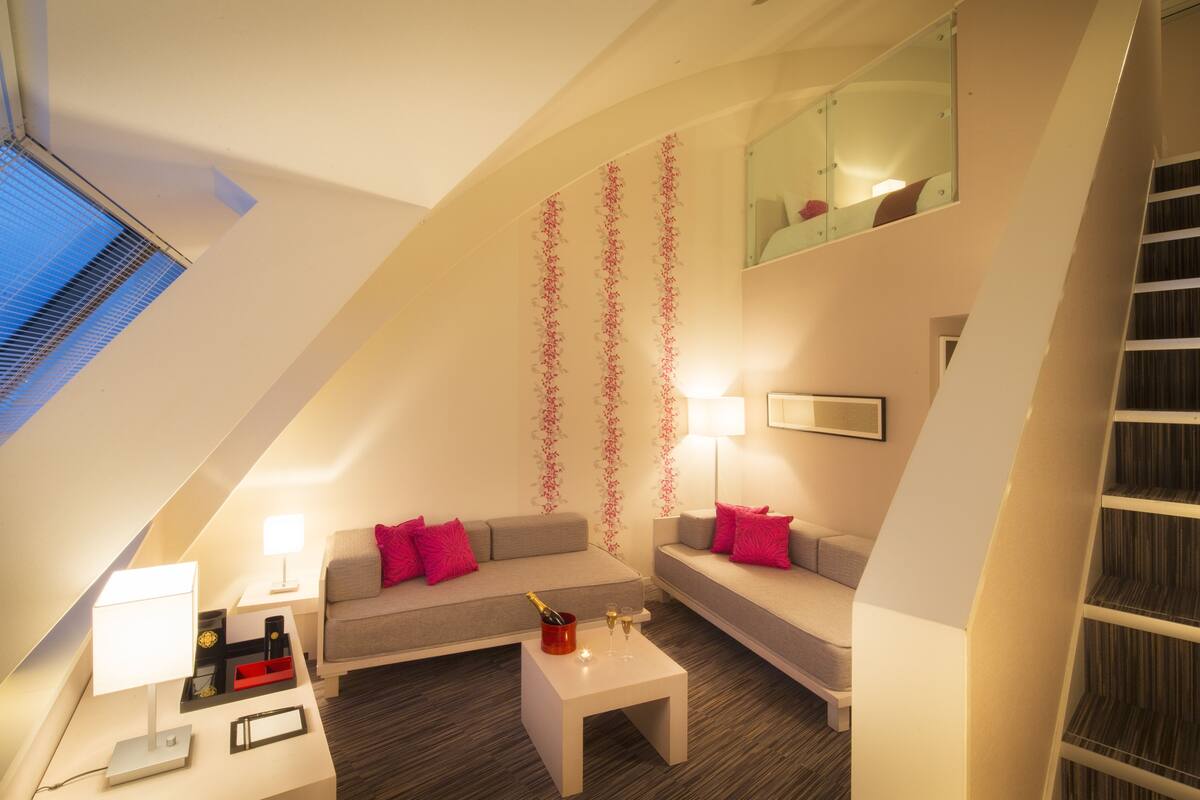 Ein stilvolles Loft-Zimmer mit schrägverdeckter Decke, einem grauen Sofa mit rosafarbenen Kissen, einem weißen Couchtisch und einem kleinen Beistelltisch mit Lampe.