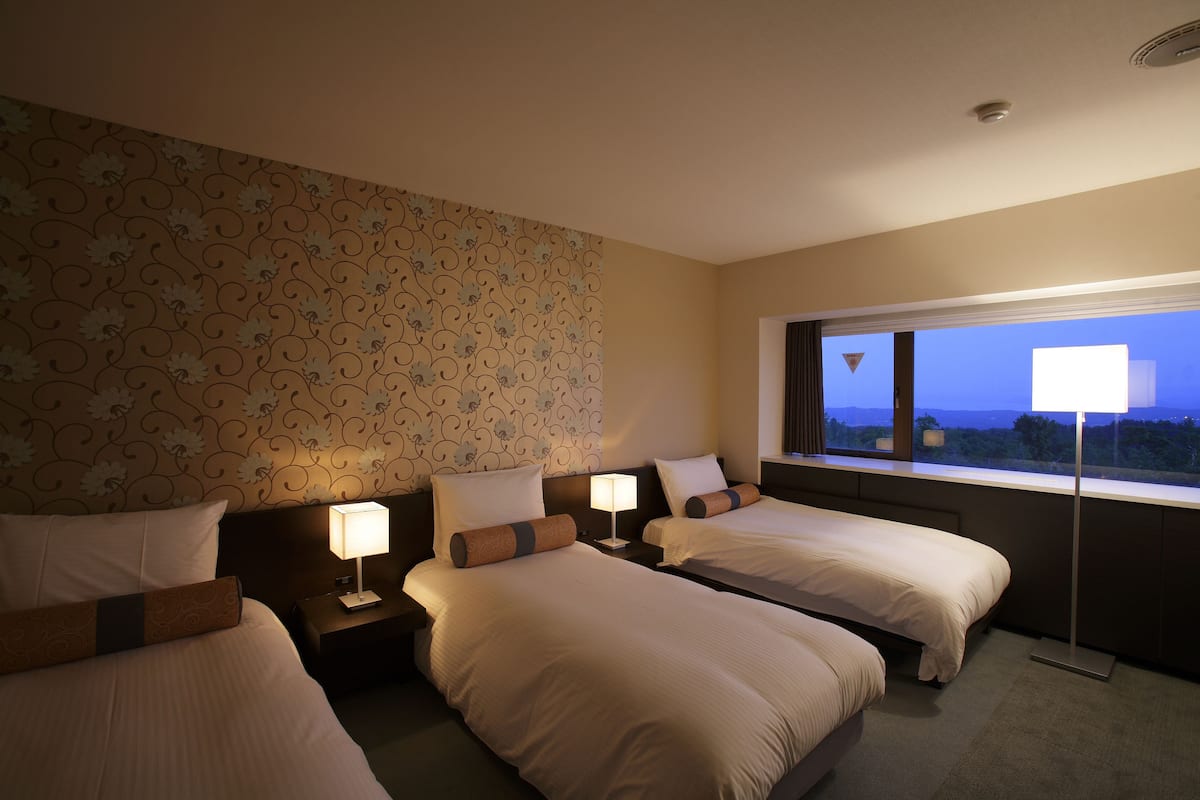 Ein Hotelzimmer mit zwei Betten, einem Nachttisch mit Lampe, einem großen Fenster mit Blick und einem Wandteppich mit Muster.