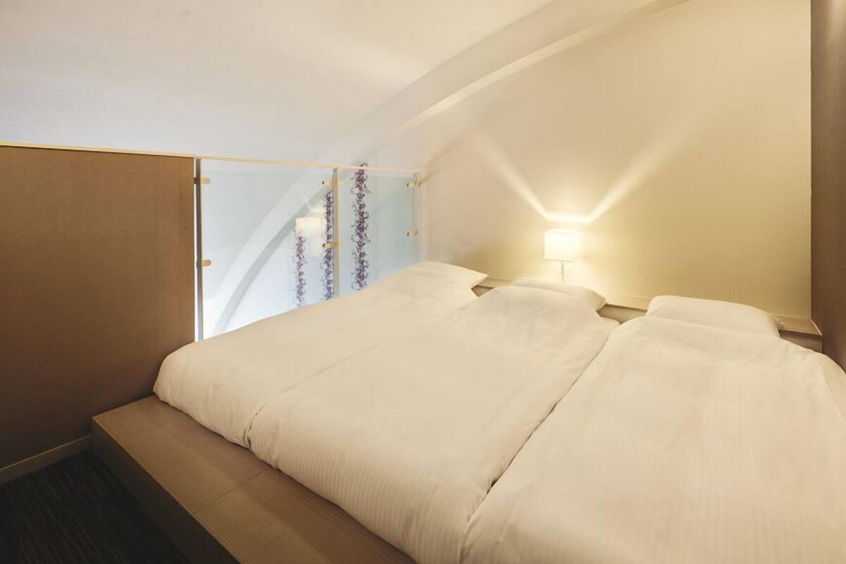 Quarto de hotel com cama, mesa de cabeceira, luminária e janela com cortinas.