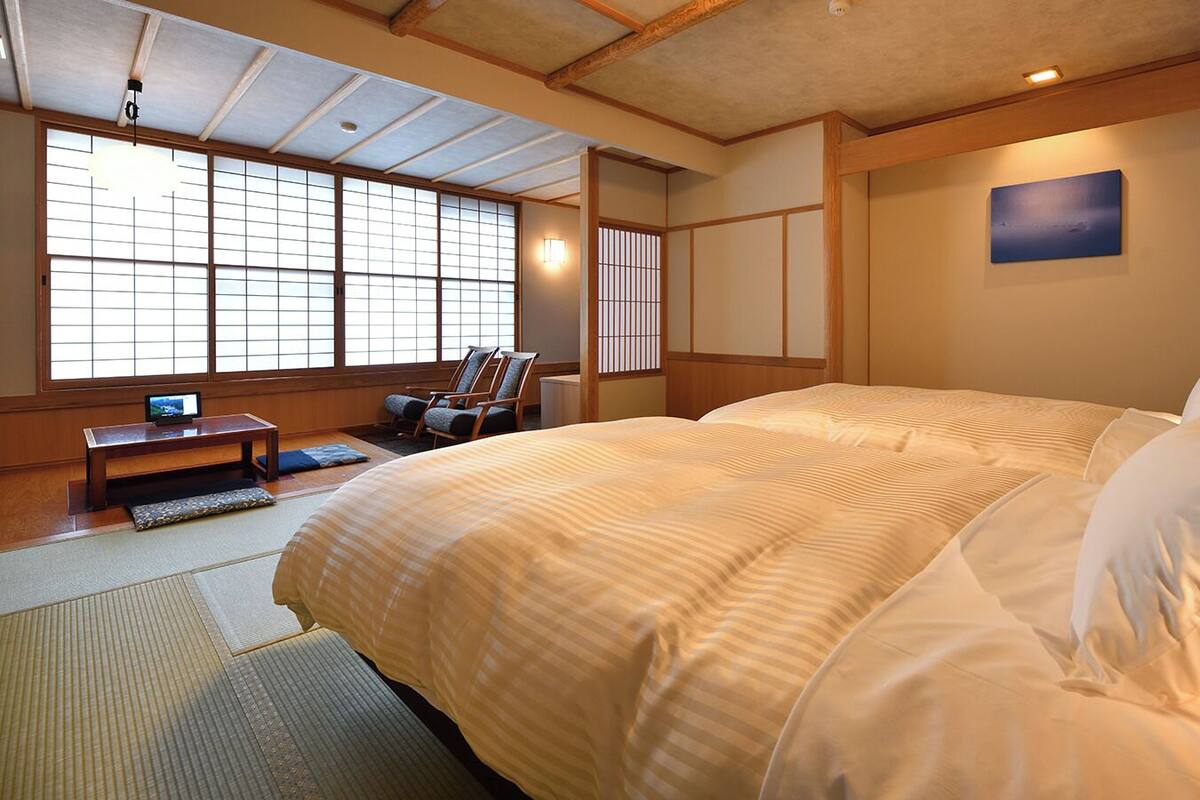 A traditional Japanese-style bedroom with tatami flooring, a large bed with white bedding, a television on a low table, and sliding doors.