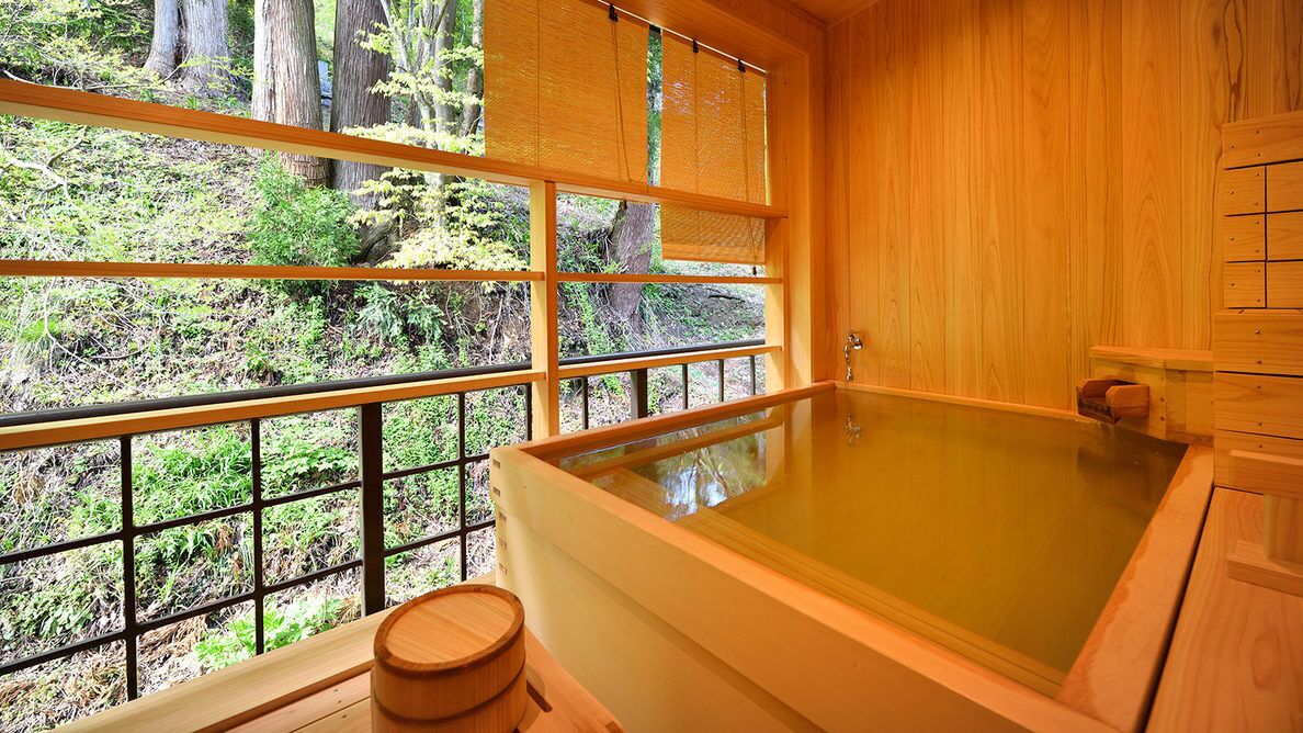A wooden room with a large bathtub, a window with a view of trees, and a wooden stool.