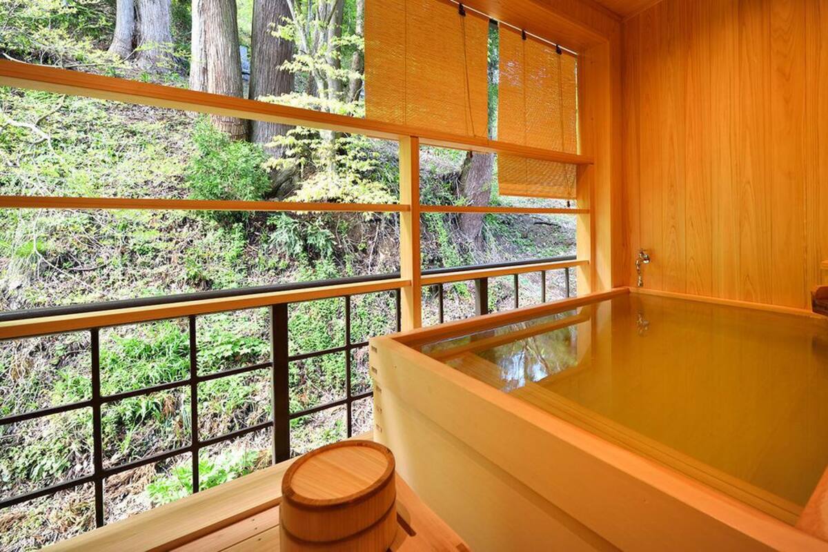 A wooden room with a large bathtub, a window with a view of trees, and a wooden stool.