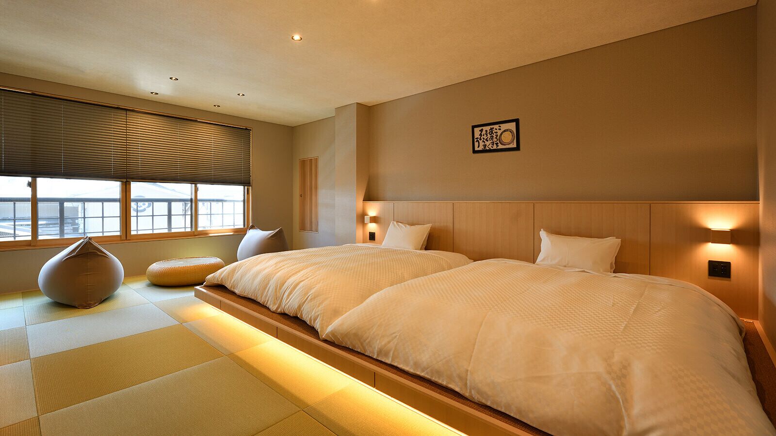 A hotel room with two beds, tatami flooring, a large window with blinds, and a wall-mounted light fixture.