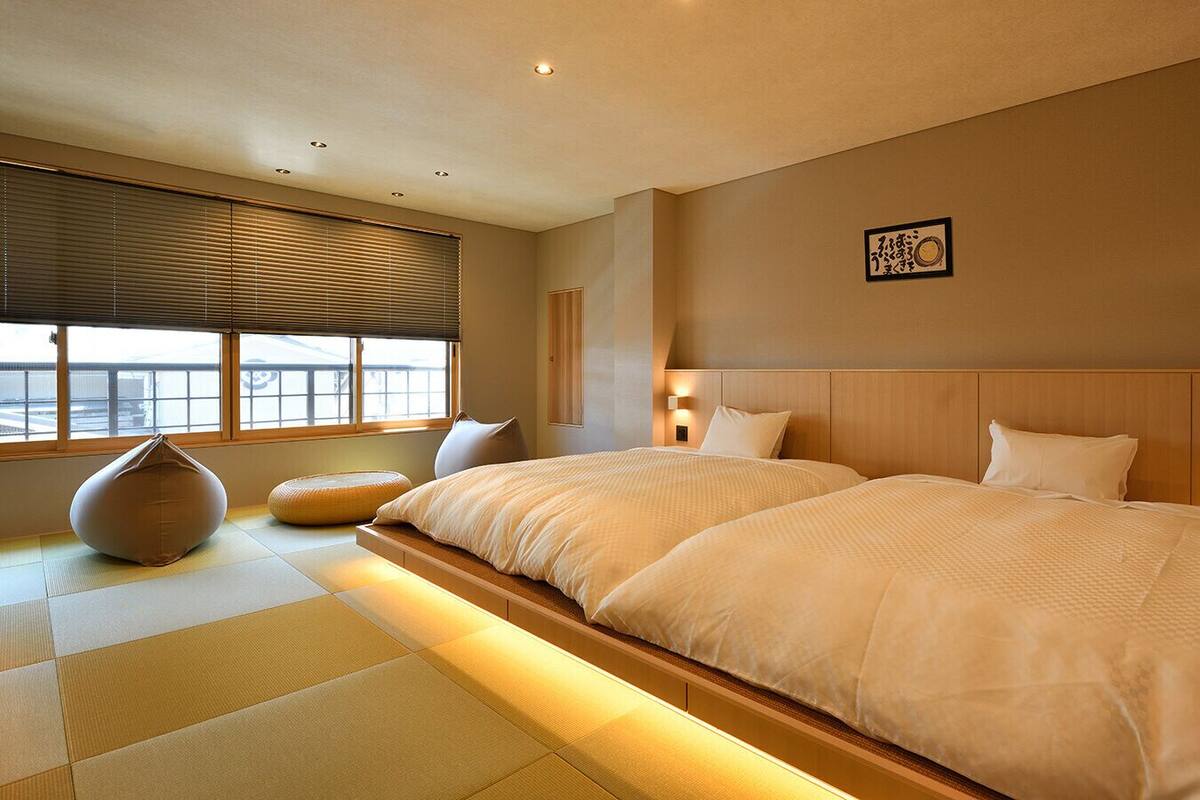 A hotel room with two beds, tatami flooring, a large window with blinds, and a wall-mounted light fixture.
