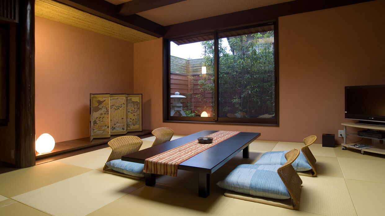 A tatami mat room with a low table, cushions, a television, and a window with a view of greenery.