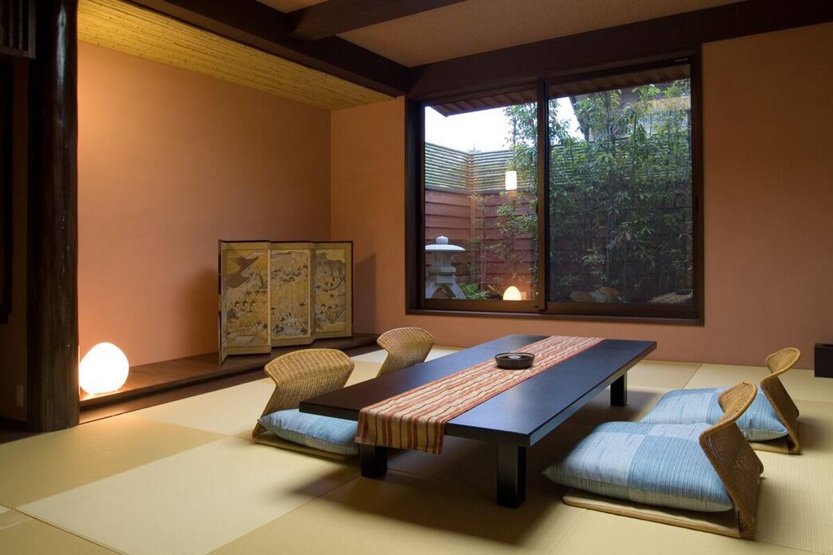 Una habitación con esteras de tatami, una mesa baja, cojines, un televisor y una ventana con vistas a la vegetación.