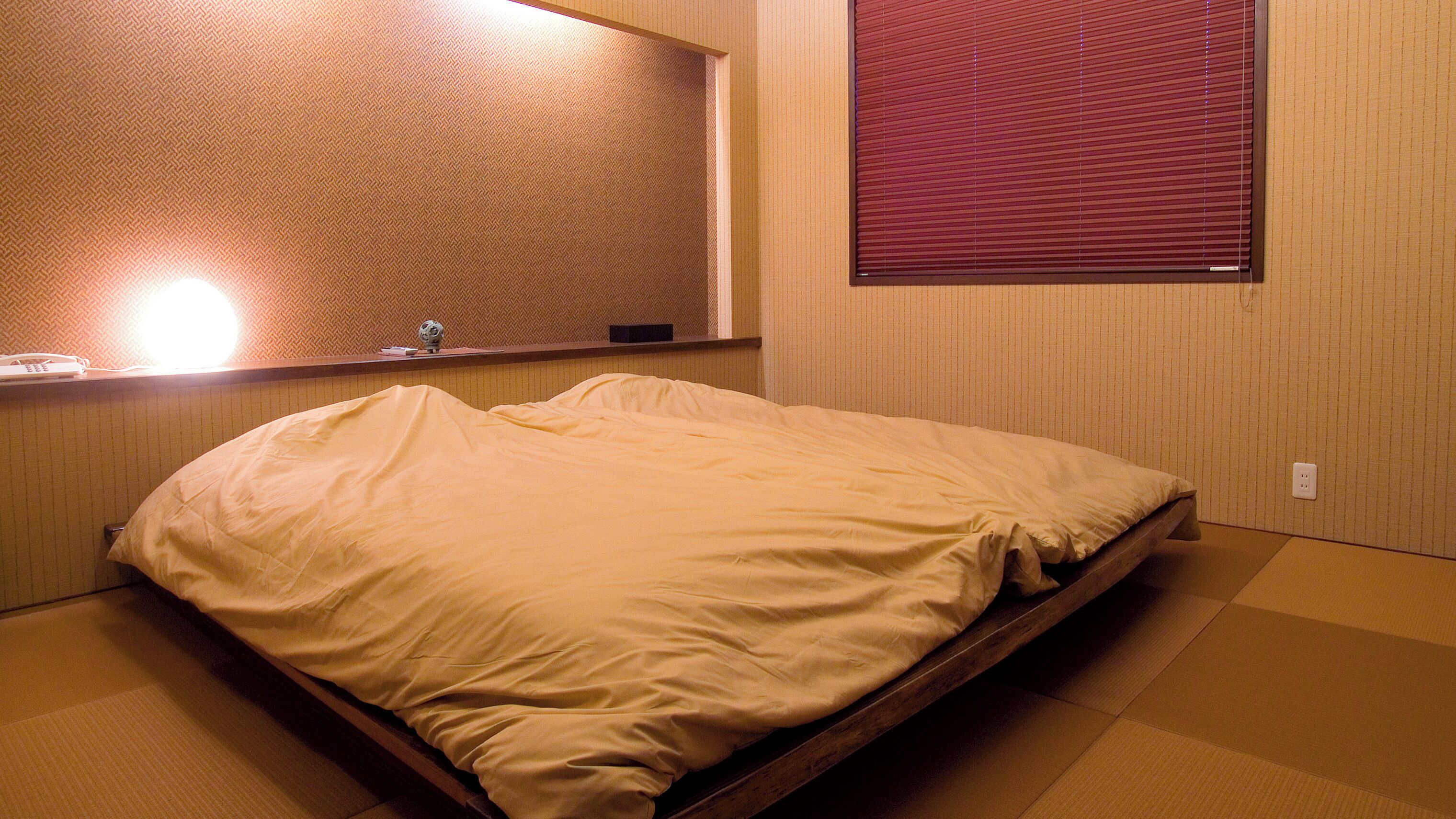 A hotel room with a bed, a bedside table, a mirror, and a window with blinds.