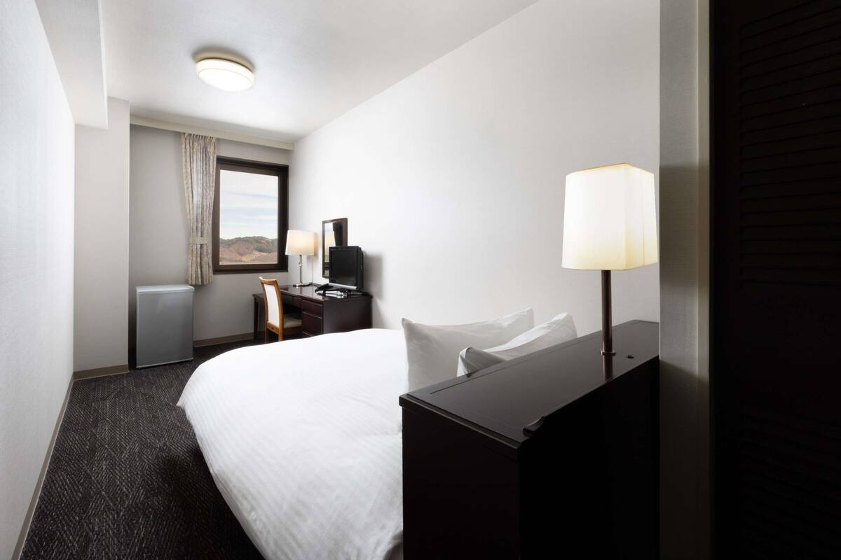 Habitación de hotel con cama, escritorio con silla, televisor, lámpara y ventana con cortinas.