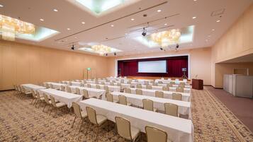A conference room with rows of tables and chairs, a stage with a red curtain, and a large screen.