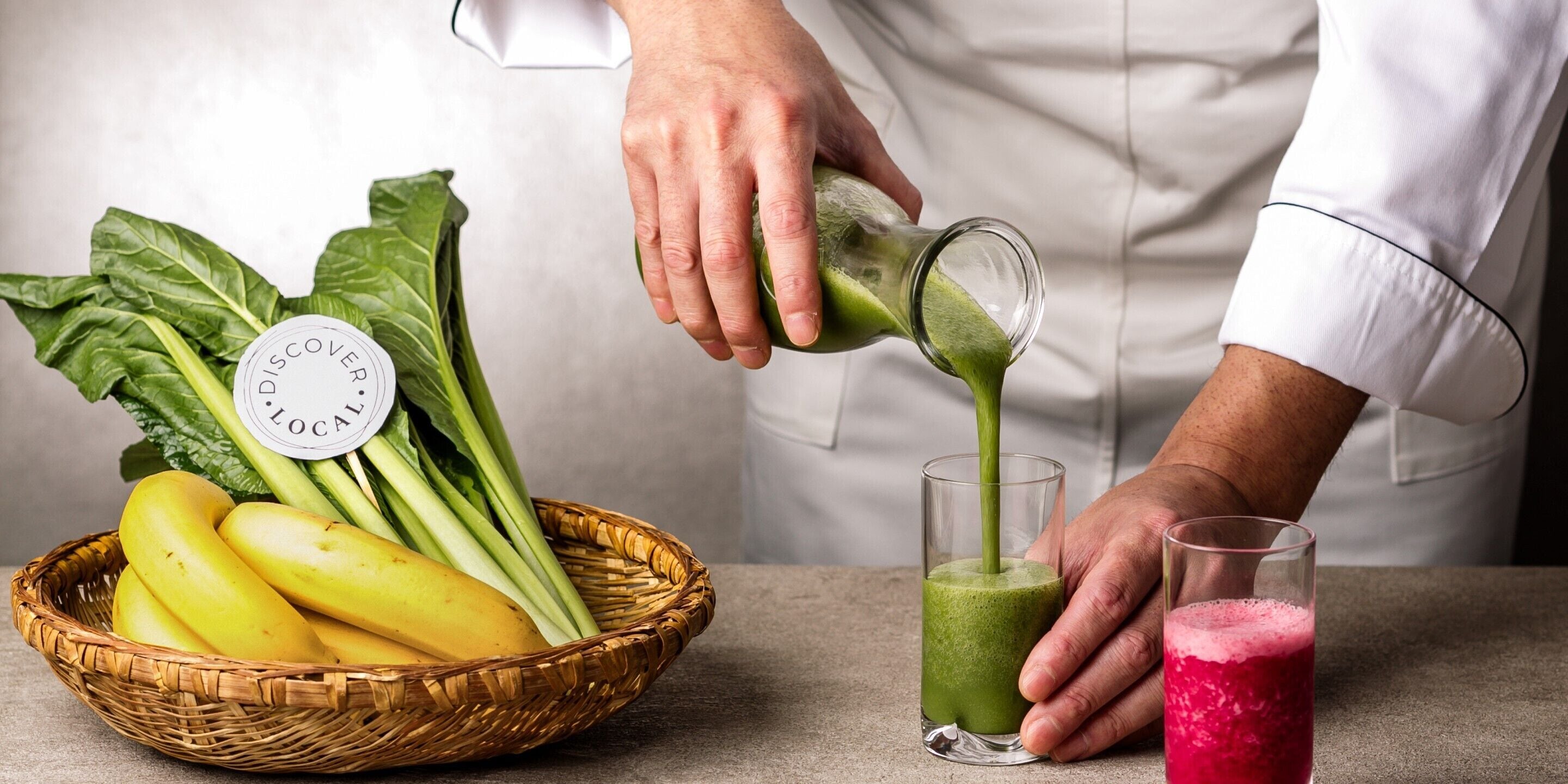 A person pouring a green smoothie into a glass, with a basket of bananas and leafy greens nearby.