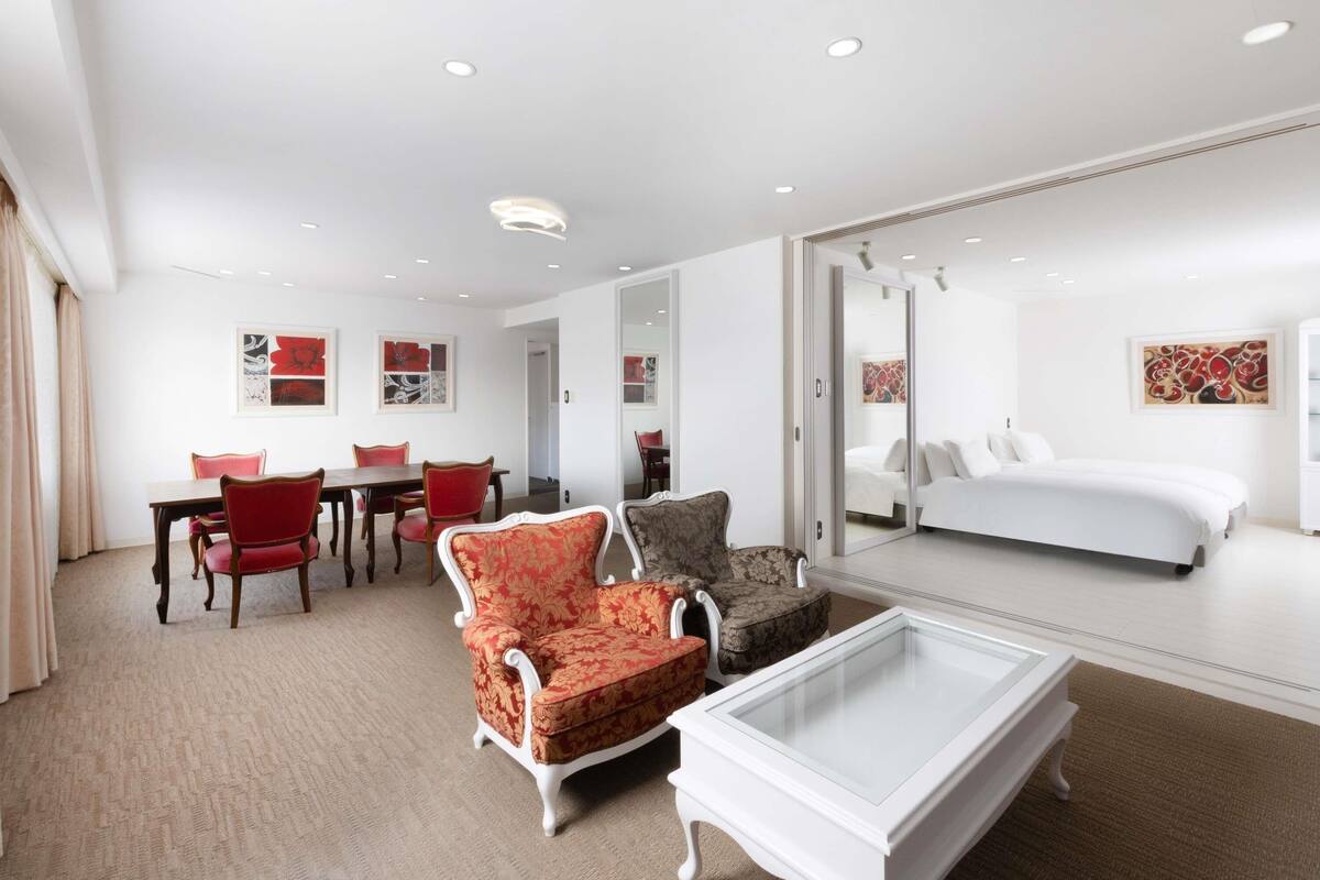 Habitación de hotel con comedor, sofá, mesa de centro de vidrio, cama y obras de arte en las paredes.