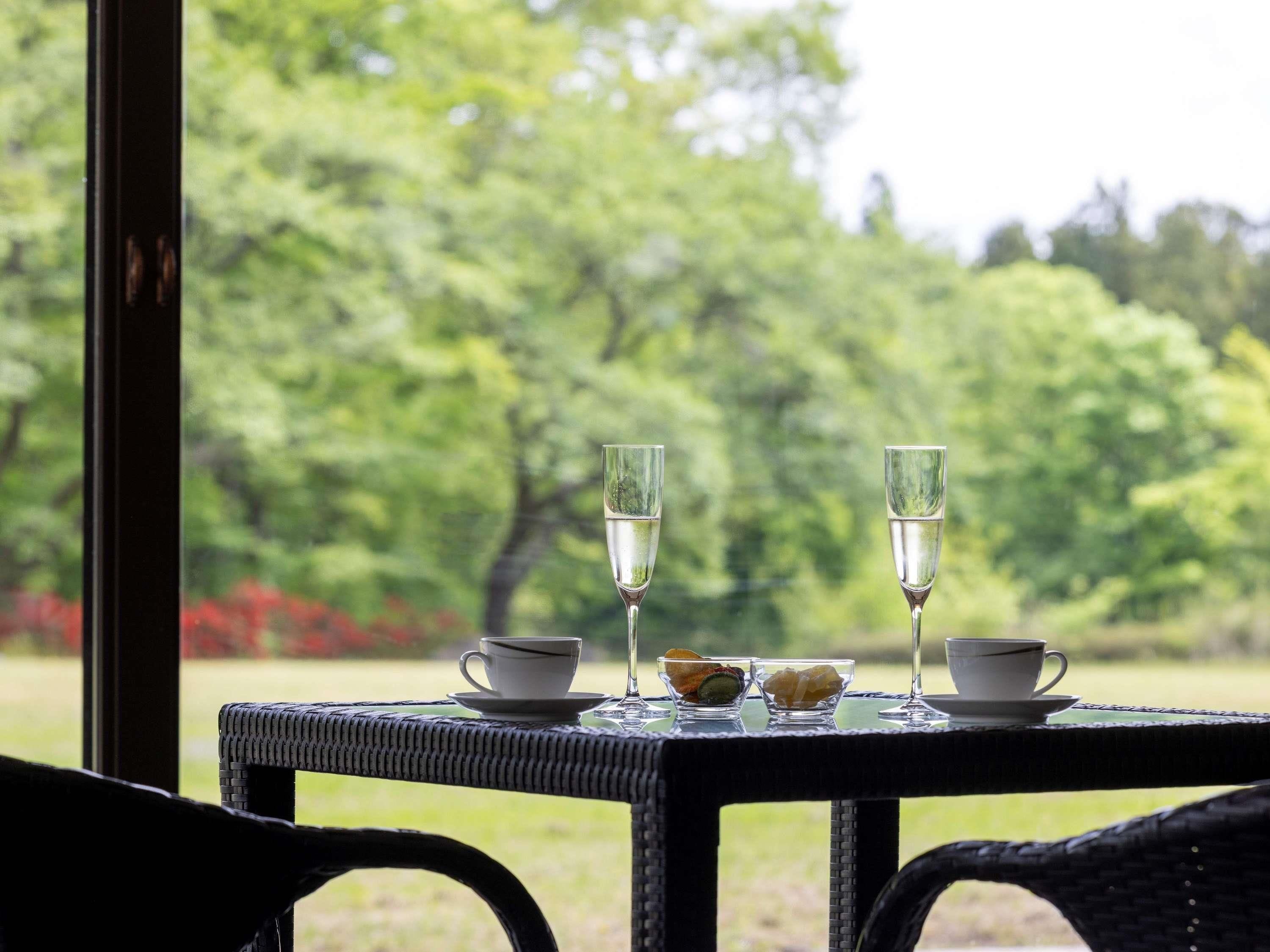 A table with two glasses of water, a cup, and a bowl of food, set against a backdrop of trees and a window.