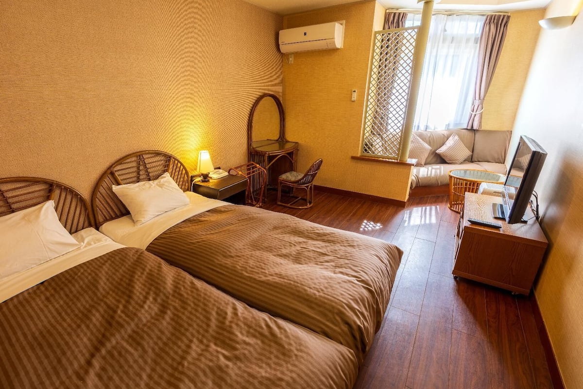 A hotel room with two beds, a desk with a lamp, a chair, a TV, and a window with curtains.